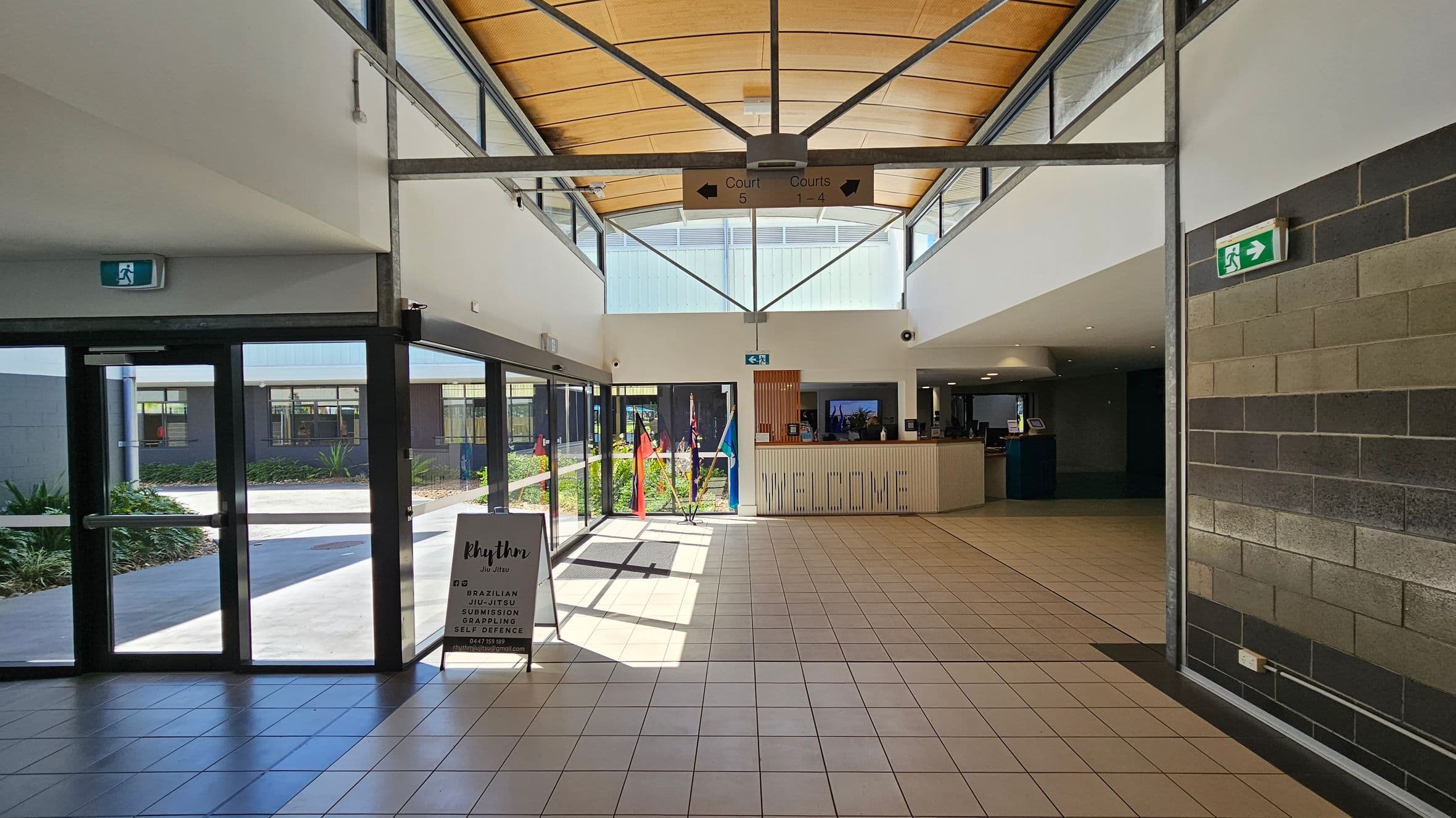 A spacious indoor area with a tiled floor and a high, wooden ceiling. On the left, there are glass doors leading outside, with a sign advertising "Rhythm Jiu Jitsu" nearby. In the centre, a reception desk with a "WELCOME" sign is visible, and flags are displayed behind it. On the right, a wall with grey and brown bricks. Above, a sign directs to courts 1-5. Emergency exit signs are positioned on both sides. Natural light streams in through large windows.