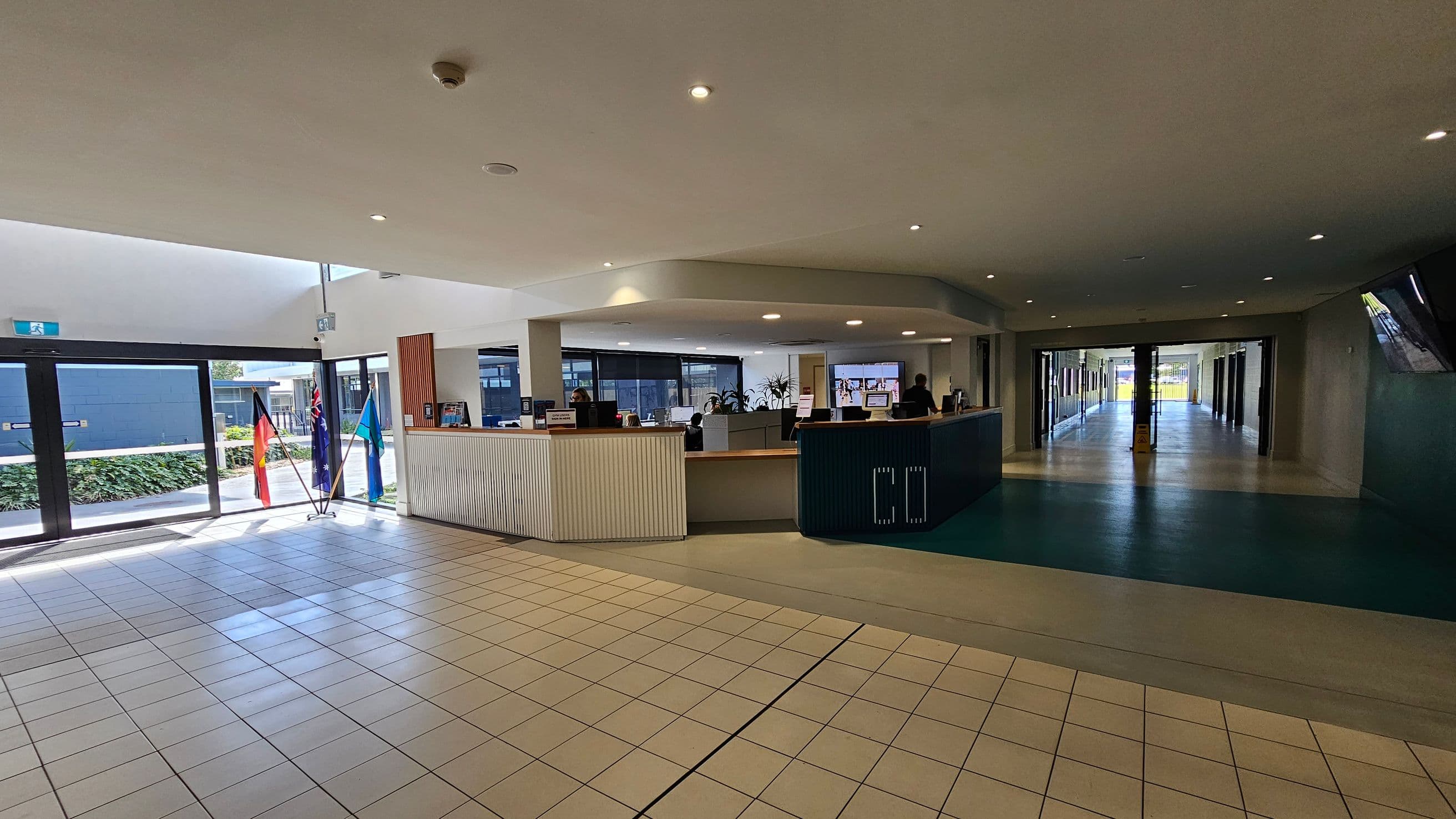 A spacious indoor area with a tiled floor and a high ceiling with recessed lighting. On the left, there are large glass doors with three flags displayed nearby. In the centre, a curved reception desk is staffed by people, with computer monitors and potted plants visible. To the right, a corridor extends into the distance, with a polished floor and glass doors at the end. The area is well-lit with natural light coming through the glass doors.