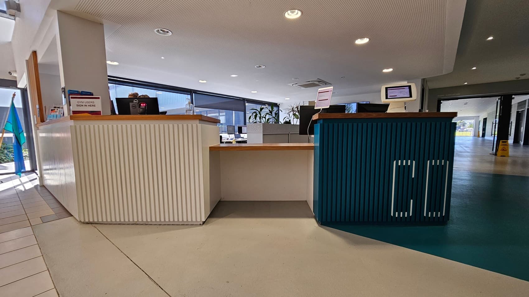 A reception area with a counter in the centre. The left side of the counter is white with a sign that reads "GYM USERS SIGN IN HERE" and some brochures. The right side is blue with a computer monitor. Behind the counter are more monitors and potted plants. The floor is a mix of light and dark tiles, and the ceiling is white with recessed lighting. Large windows in the background let in natural light.