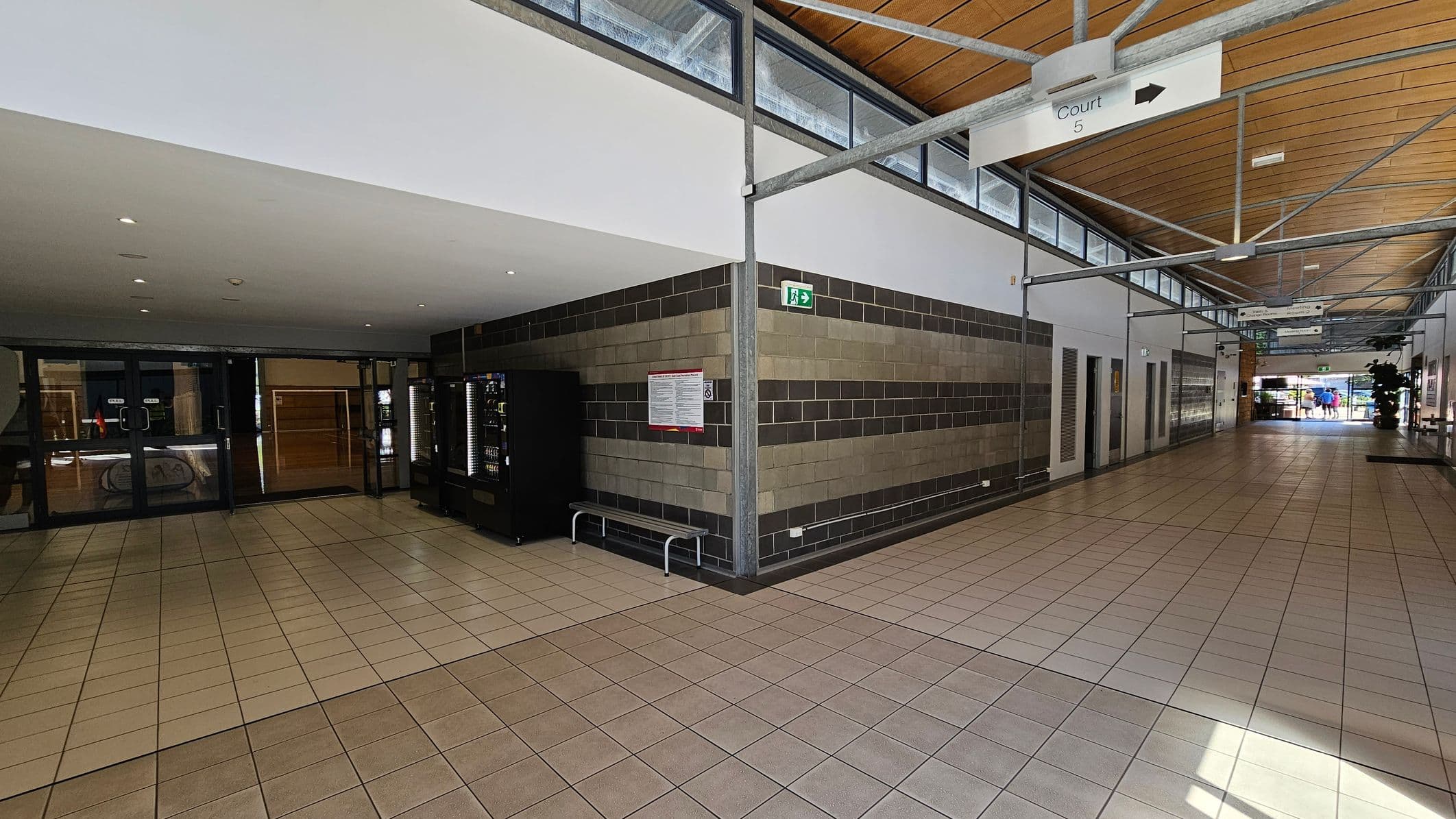 A corridor with a tiled floor and a wooden panelled ceiling. On the left, there are glass doors leading to another area, with vending machines nearby. The right side features a wall with a striped pattern of grey and beige bricks, a bench, and an emergency exit sign. Further down the corridor, signs hang from the ceiling, and there is a glimpse of people near the entrance at the far end.