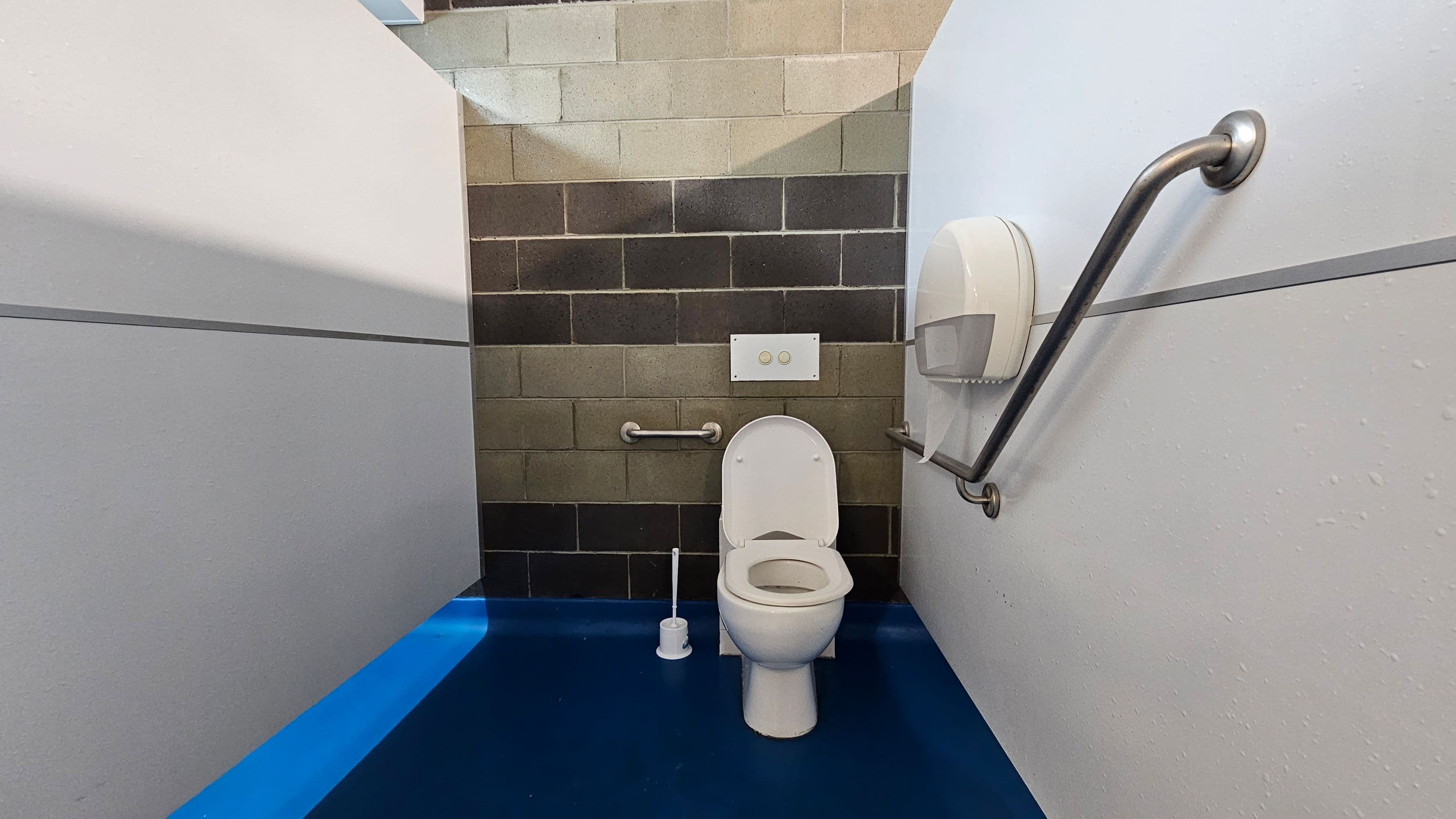 A public toilet with a white toilet bowl and seat in the centre. The back wall is made of multicoloured bricks. There are two metal grab bars on either side of the toilet, with a toilet paper dispenser on the right. The floor is a bright blue, and the walls are white with a horizontal dividing line. A toilet brush is placed on the floor to the left of the toilet.