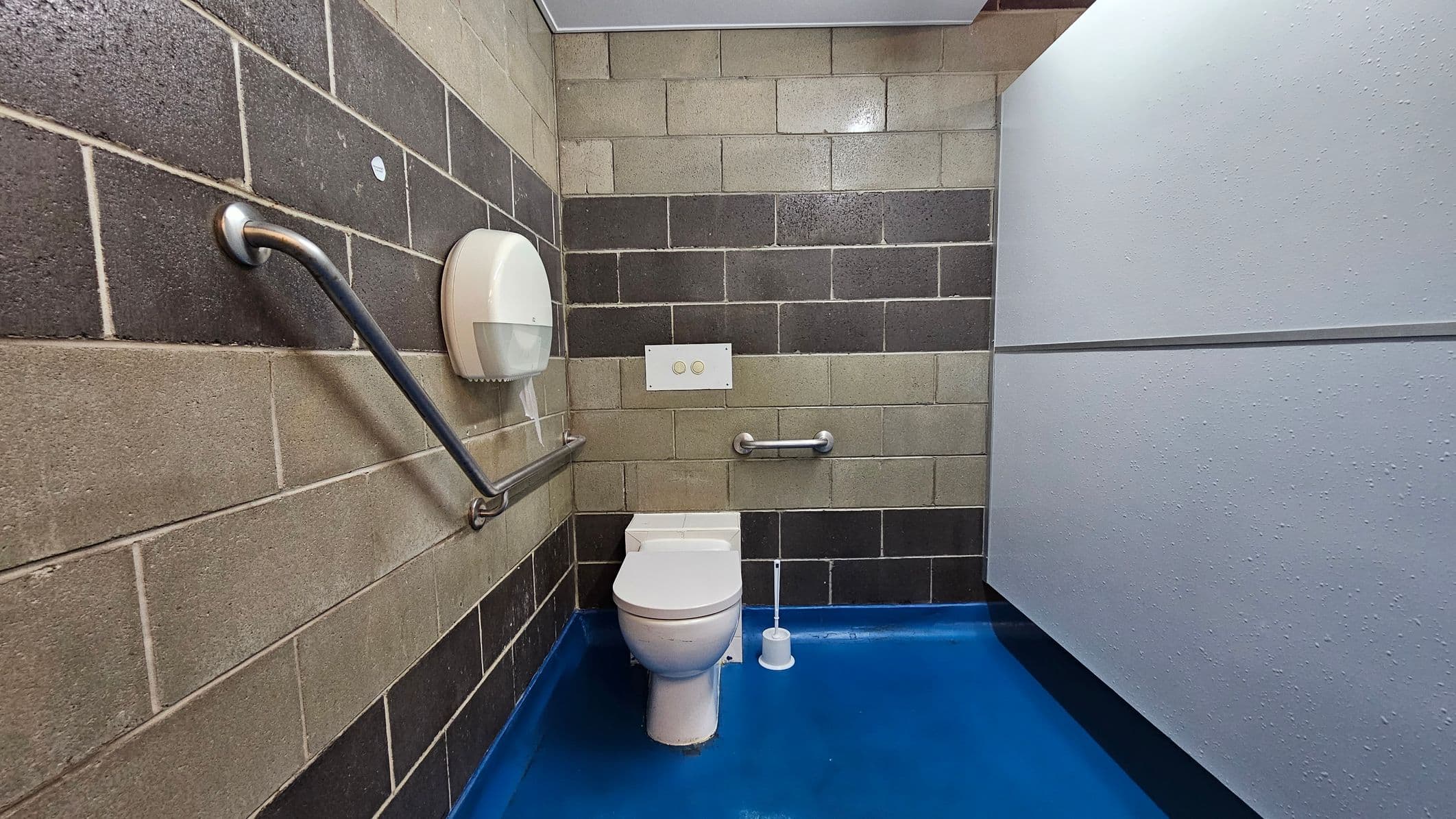 A public toilet with a blue floor and a white ceiling. The walls are made of grey and beige concrete blocks. On the left, there's a metal grab bar and a toilet paper dispenser. In the centre, a white toilet with a dual-flush button panel above it and another grab bar to the right. A toilet brush is placed on the floor next to the toilet. On the right, a light grey partition wall.