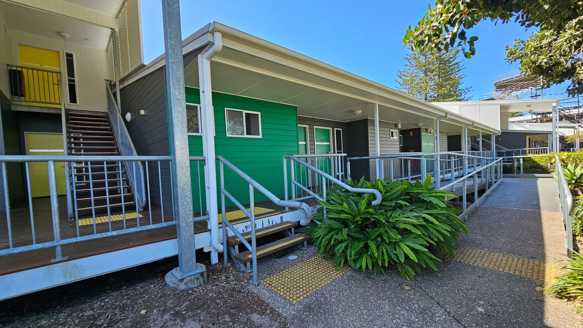A building with a green exterior wall and a covered walkway. On the left, there is a staircase with a metal railing leading to an upper level with a yellow door. The foreground features a ramp with handrails and tactile paving. A lush green plant is near the ramp. The building has a flat roof and a grey section further along. Trees and a structure with a metal framework are visible in the background. The ground is paved with a textured surface.