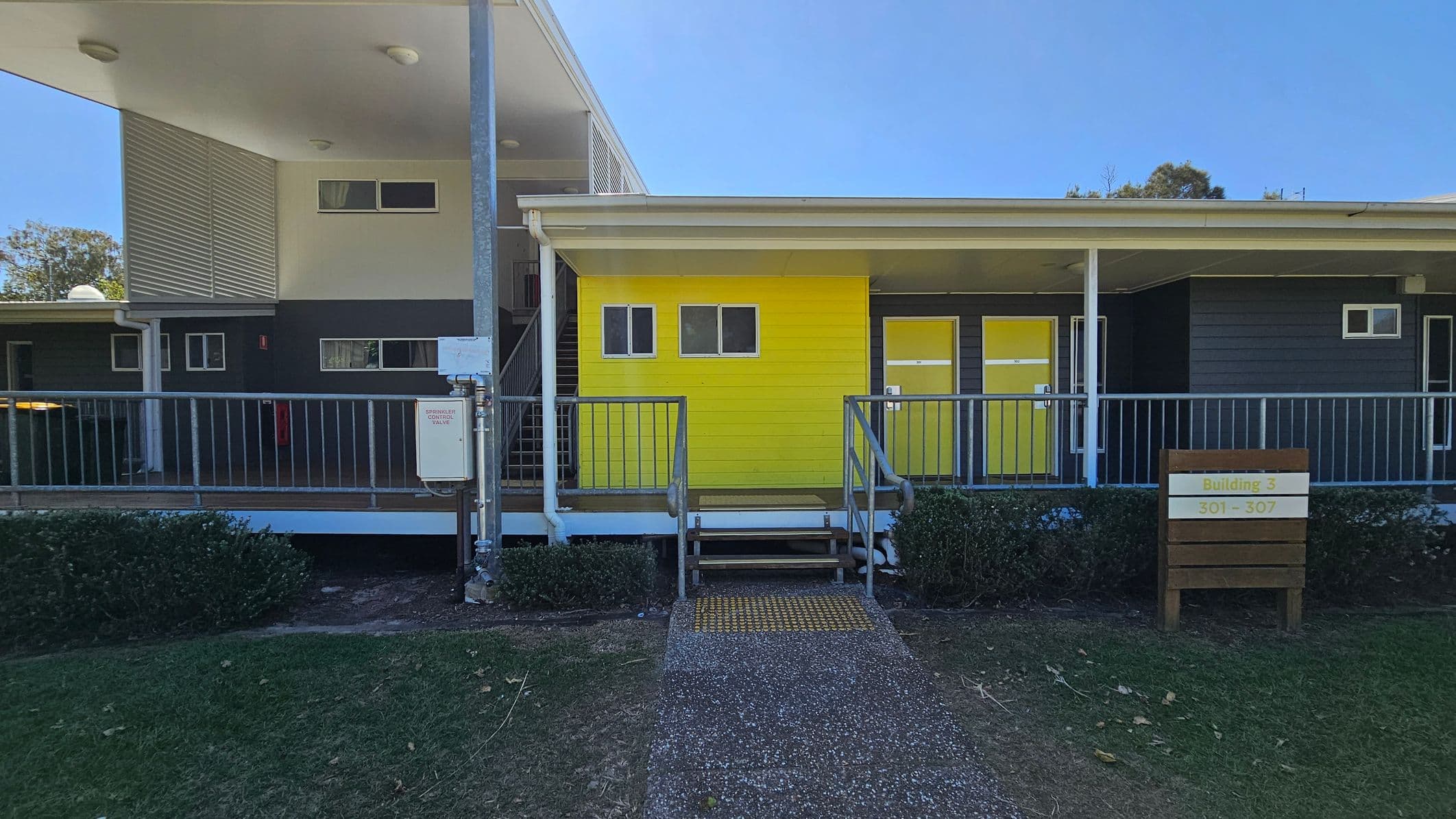 A building with a bright yellow section in the centre, featuring two small windows and a door. To the right, there is a sign reading "Building 3, 301 - 307" and another yellow door. The left side has a grey section with a staircase and additional windows. A metal railing runs along the front, with a small set of stairs leading up to the entrance. The foreground has a path and green grass, with bushes lining the building. The ceiling is white with recessed lights.