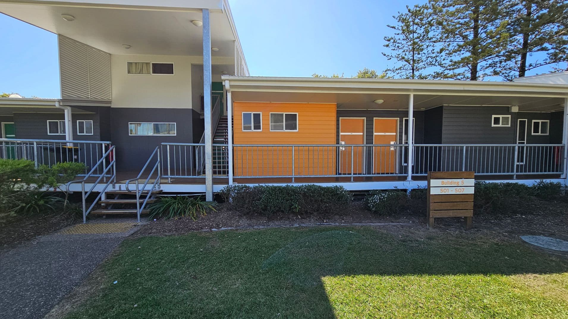 A modern building with a flat roof and a mix of grey and orange panelling. On the left, there is a staircase leading to an upper level. The building has several small rectangular windows. In front, a metal railing runs along a walkway. A wooden sign in the foreground reads "Building 5, 501 - 507". The ground is covered with grass and shrubs, and tall trees are visible in the background.