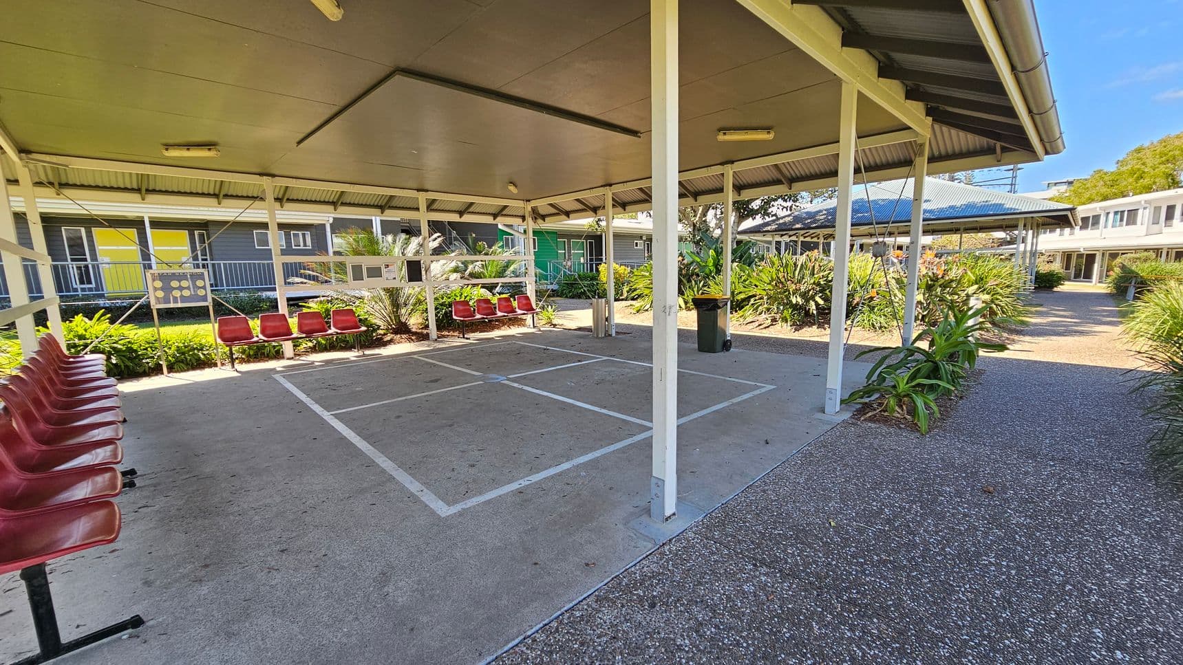 A covered outdoor area with a concrete floor marked with white lines. Red plastic chairs are lined up on the left side. The structure has a metal roof supported by white poles. Surrounding the area are green plants and shrubs. In the background, there are buildings with yellow and green walls, and a pathway with a speckled surface leads through the space. A rubbish bin is visible near the centre.