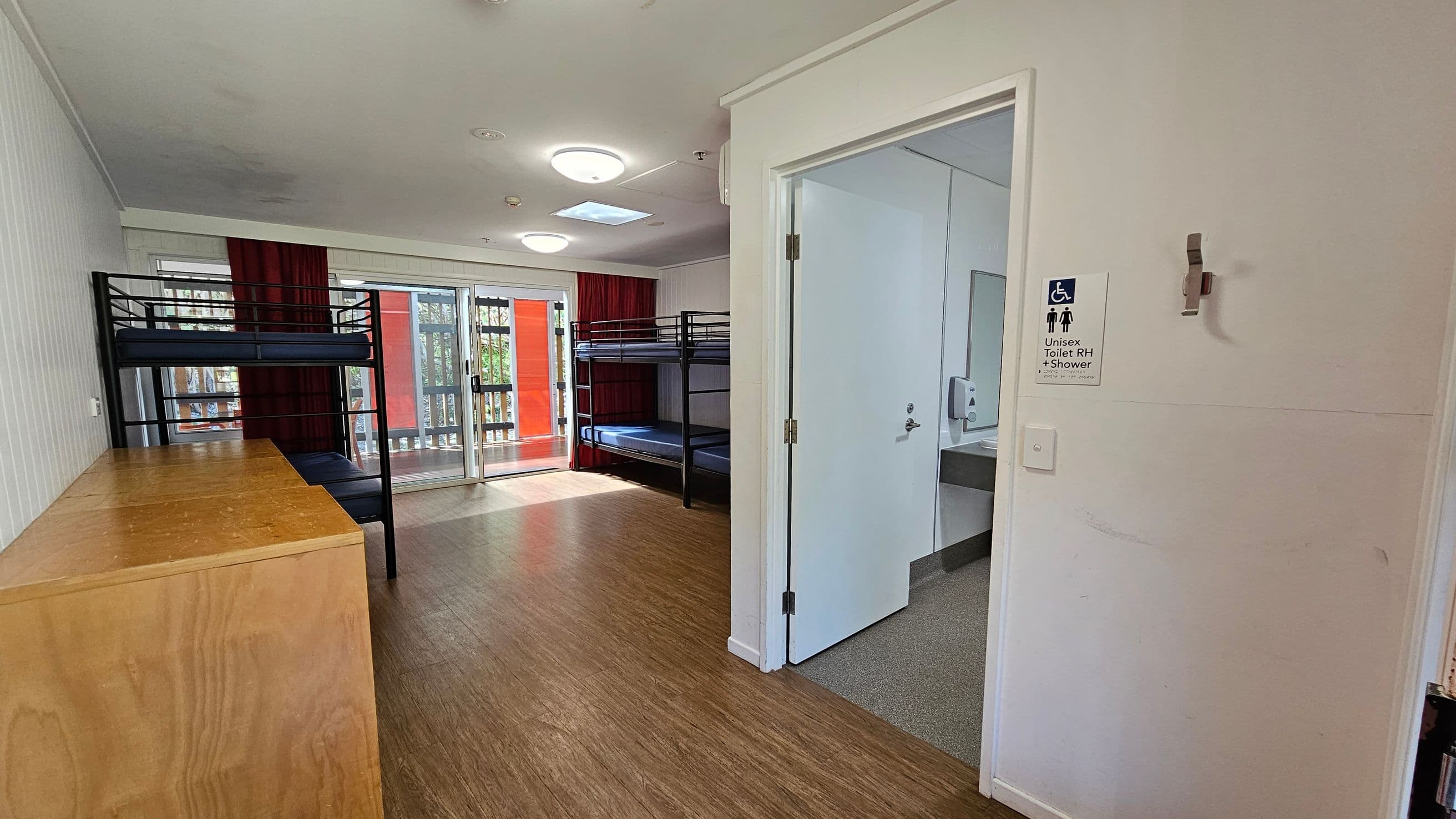 A room with wooden flooring and a white ceiling. On the left, there are two sets of metal bunk beds and a wooden cabinet. In the centre, a sliding glass door with red curtains leads to a balcony. On the right, an open door reveals a bathroom with a sign indicating it is a unisex toilet with a shower. The bathroom has a sink and a soap dispenser visible.