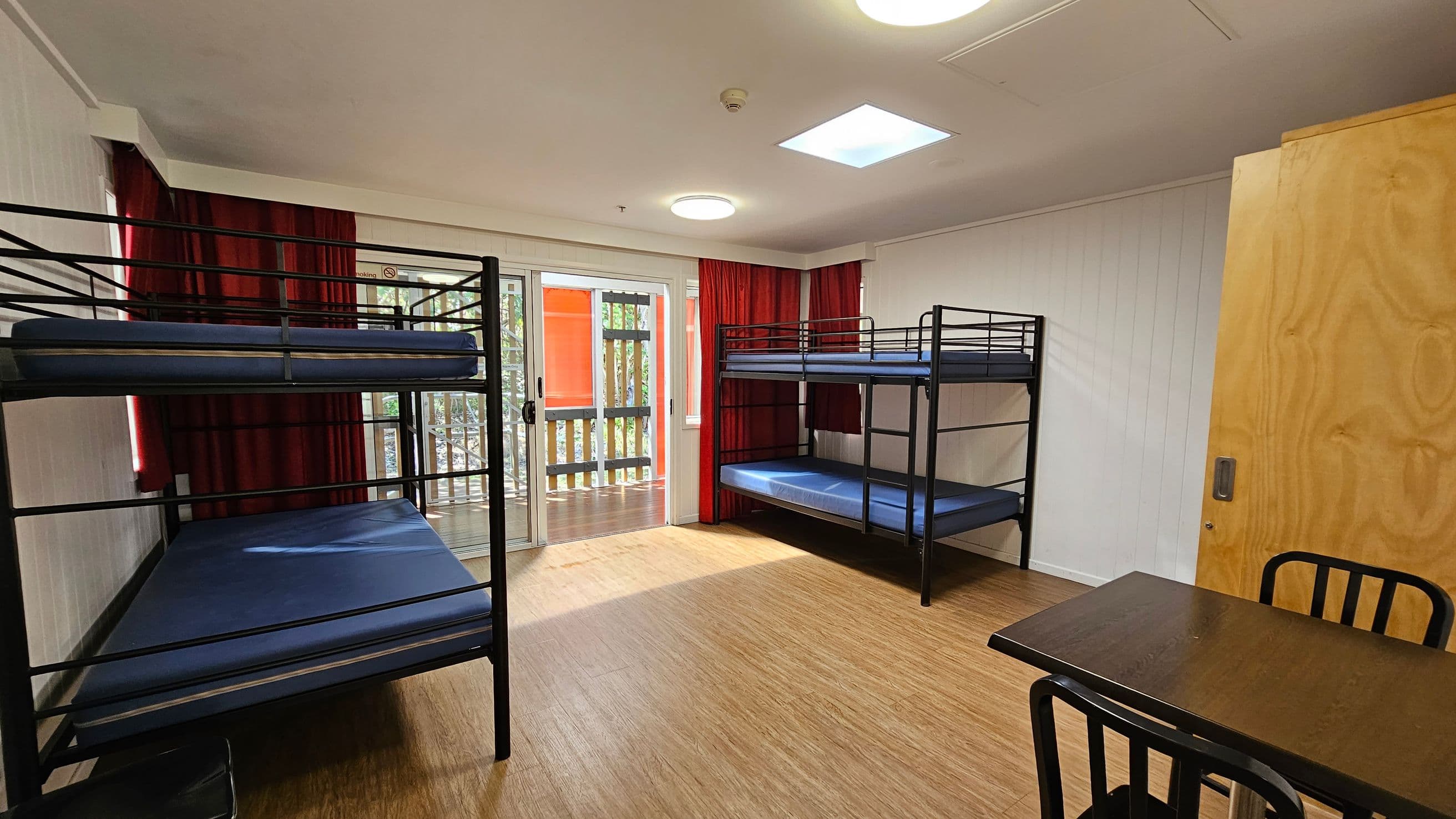 A room with two sets of black metal bunk beds, each with blue mattresses, positioned on the left and right sides. In the centre, there are large sliding glass doors with red curtains, leading to a balcony with a wooden railing. On the right, a wooden door and a small black table with chairs are visible. The floor is light wood, and the ceiling has recessed lighting and a skylight.