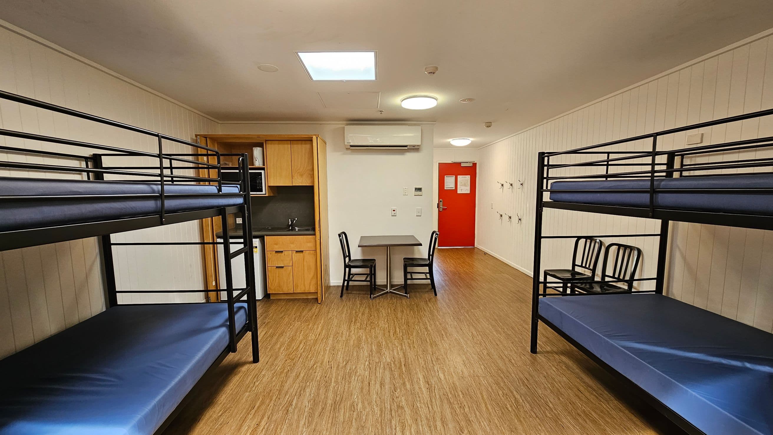 A room with two sets of metal bunk beds on the left and right, each with blue mattresses. In the centre, there's a small table with two black chairs. The back wall features a red door and a wall-mounted air conditioning unit above the table. To the left, there's a kitchenette with wooden cabinets, a microwave, and a small sink. The floor is wooden, and the walls are white with vertical panelling. Ceiling lights are visible above.
