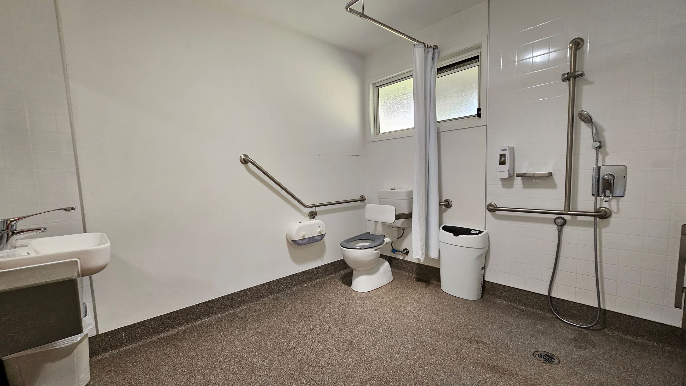 A spacious, accessible bathroom with a non-slip brown floor and light-coloured walls. On the left, there's a sink with a single lever tap. In the centre, a toilet with grab bars on both sides and a toilet paper holder. To the right, a shower area with a handheld showerhead and additional grab bars. A white curtain separates the shower from the rest of the room. A small window is above the toilet, and a waste bin is nearby.
