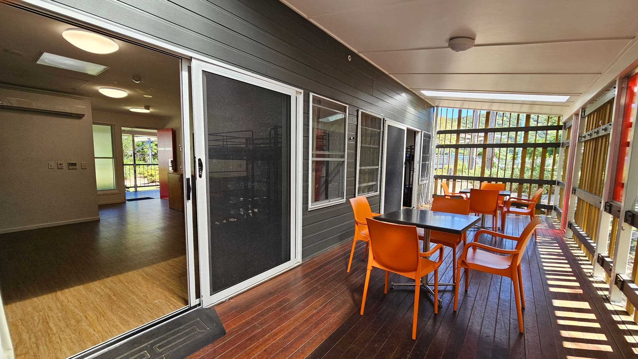 A covered outdoor area with wooden flooring and a ceiling with recessed lighting. On the right, there is a table with four bright orange chairs. The left side features sliding glass doors leading to an interior space with wooden flooring and ceiling lights. The background shows vertical wooden slats allowing natural light to filter through, with greenery visible beyond.