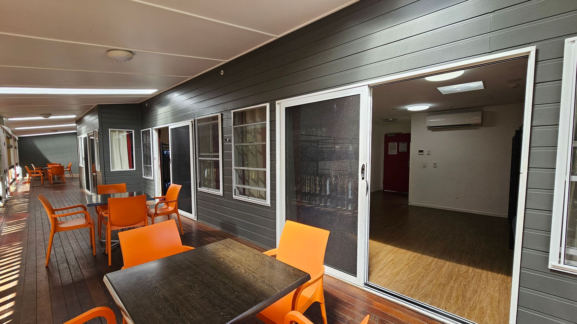 A covered outdoor area with a wooden floor and a white ceiling. On the left, there are several tables with bright orange chairs. The right side features a dark grey wall with multiple windows and a sliding glass door leading to an interior room. The room inside has a wooden floor, white walls, and a visible air conditioning unit. The space is well-lit with ceiling lights.