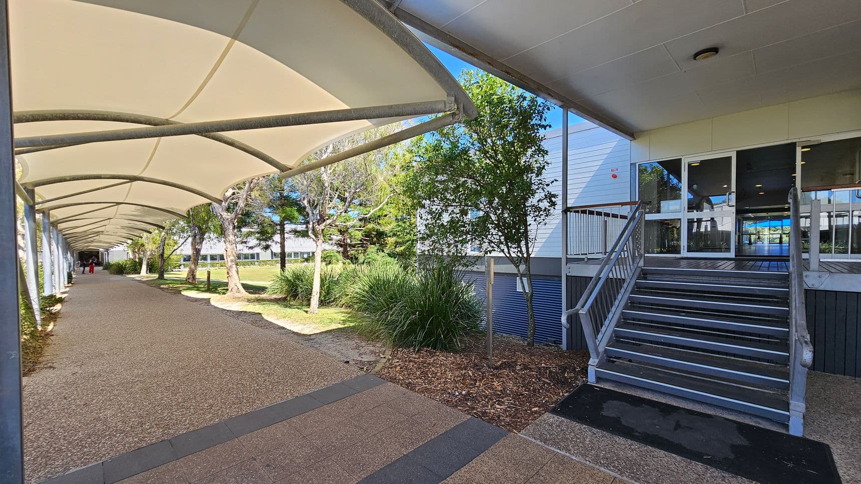 A covered walkway on the left with a curved, white canopy supported by metal beams. The path is paved with a textured surface. On the right, there is a building with a set of metal stairs leading to glass doors. Trees and shrubs are planted along the side of the building, and a grassy area is visible in the background. The ceiling of the walkway is made of fabric, and the building has a flat, white ceiling.