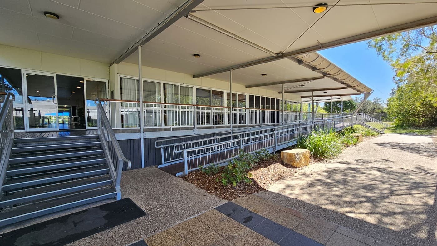 A building entrance with a covered walkway. On the left, there are metal stairs leading up to glass doors. The middle section features a long ramp with railings, bordered by a small garden with plants and a large stone. On the right, a paved path extends alongside the building, surrounded by trees and greenery. The ceiling is flat with recessed lights, and the floor is a mix of textured and smooth surfaces.