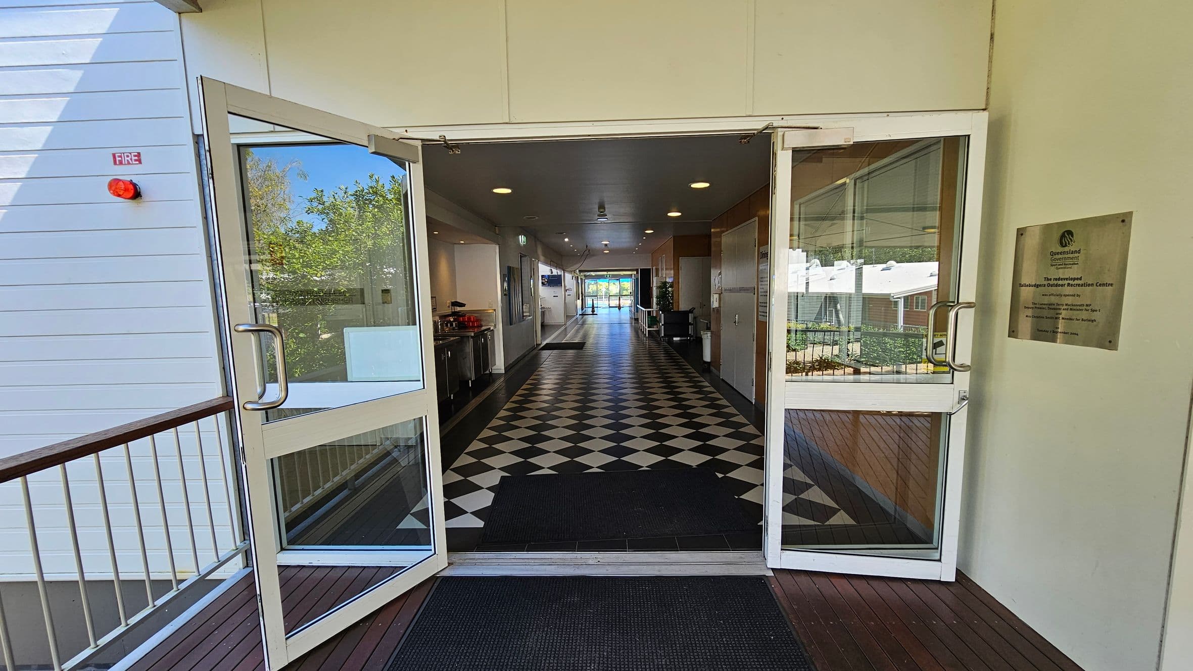 Glass double doors open into a long corridor with a black and white chequered floor. On the left, there's a small kitchen area with a counter and appliances. On the right, a plaque is mounted on the wall. The corridor is well-lit with ceiling lights and leads to a bright area with large windows at the far end. Outside the doors, a wooden deck with metal railings is visible, along with some greenery.
