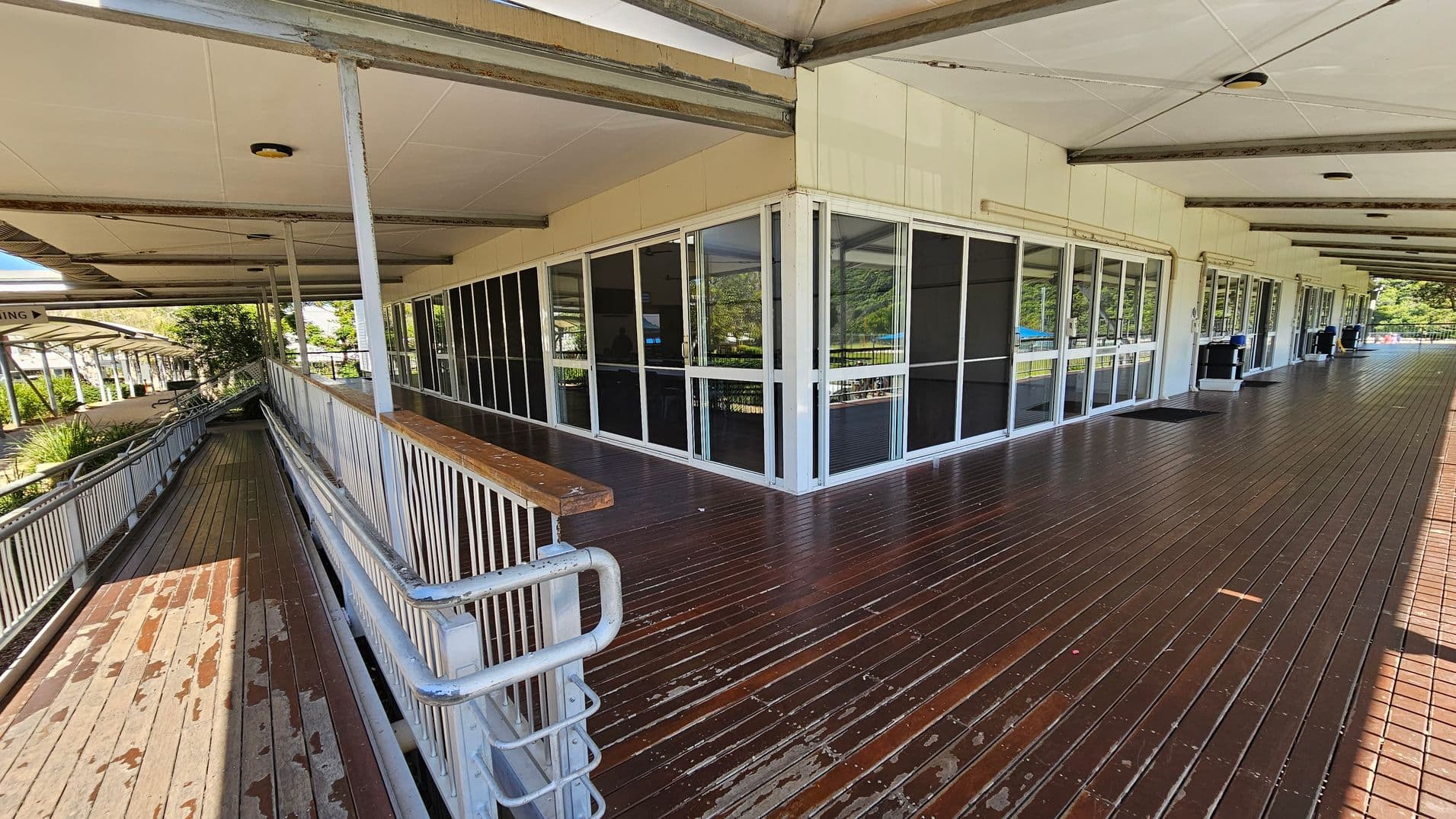 A wooden deck with a ramp on the left side, featuring metal railings. The building has large glass windows and doors with dark panels. The ceiling is supported by metal beams, and there are several ceiling lights. In the background, there are trees and a covered walkway. The floor is made of dark wooden planks, and there are a few scattered items on it.