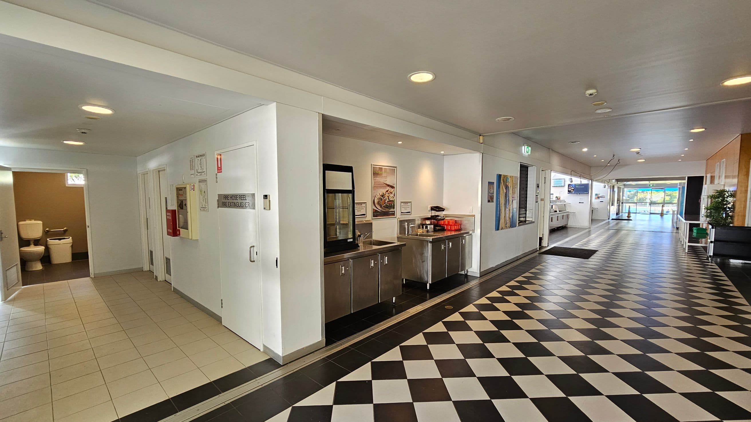 A corridor with a black and white chequered floor. On the left, there is a tiled area leading to a bathroom with visible toilets. Adjacent to this is a wall with a fire hose reel and extinguisher cabinet. In the middle, a stainless steel counter with a sink and red trays is set against the wall. The right side of the corridor has a series of artworks and leads to a glass door with a view outside. The ceiling is white with recessed lighting.