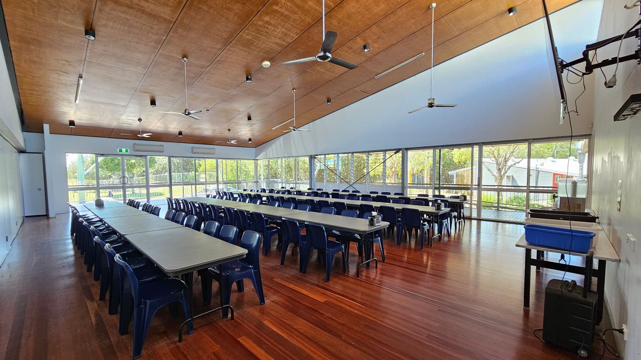 A spacious room with wooden flooring and a wooden ceiling featuring ceiling fans and lights. Long tables with blue chairs are arranged in rows, filling the room. Large windows on the right side let in natural light and offer a view of trees and a building outside. On the right, a table holds a blue container and some equipment. The room has a clean, organised appearance, suitable for meetings or dining.