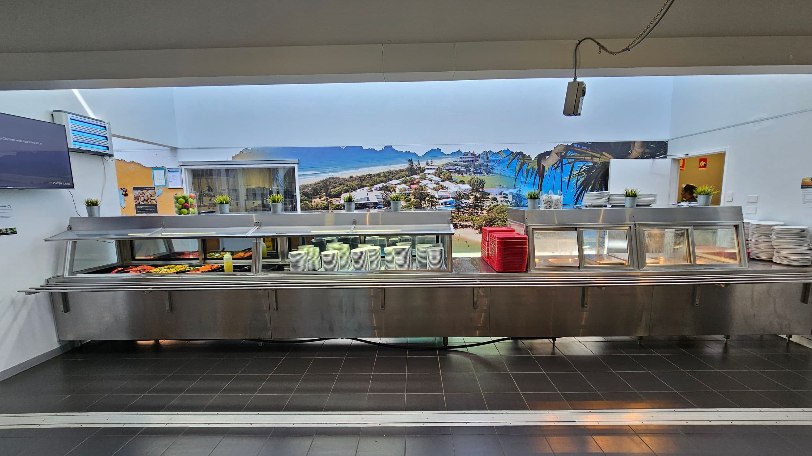 A cafeteria counter with a stainless steel serving area. On the left, there are trays of food, including vegetables and a bottle of sauce. In the centre, stacks of white plates and red trays are visible. On the right, more plates are stacked. Above the counter, small potted plants are placed. The background features a large mural of a coastal landscape with buildings and trees. The floor is tiled in dark grey, and a TV screen is mounted on the left wall.