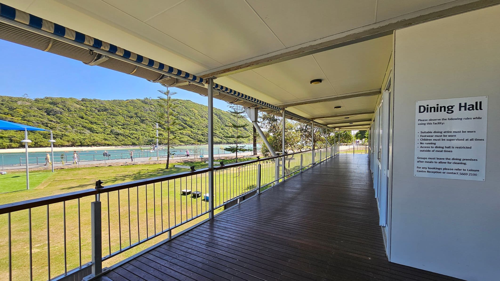 A covered walkway with a wooden floor and a white ceiling runs along the right side, featuring a sign for the Dining Hall with rules. On the left, a railing overlooks a grassy area with a few trees and a blue sunshade. Beyond the grass, there's a body of water with a fence and people walking nearby. In the background, a lush, green hill rises, providing a natural backdrop.
