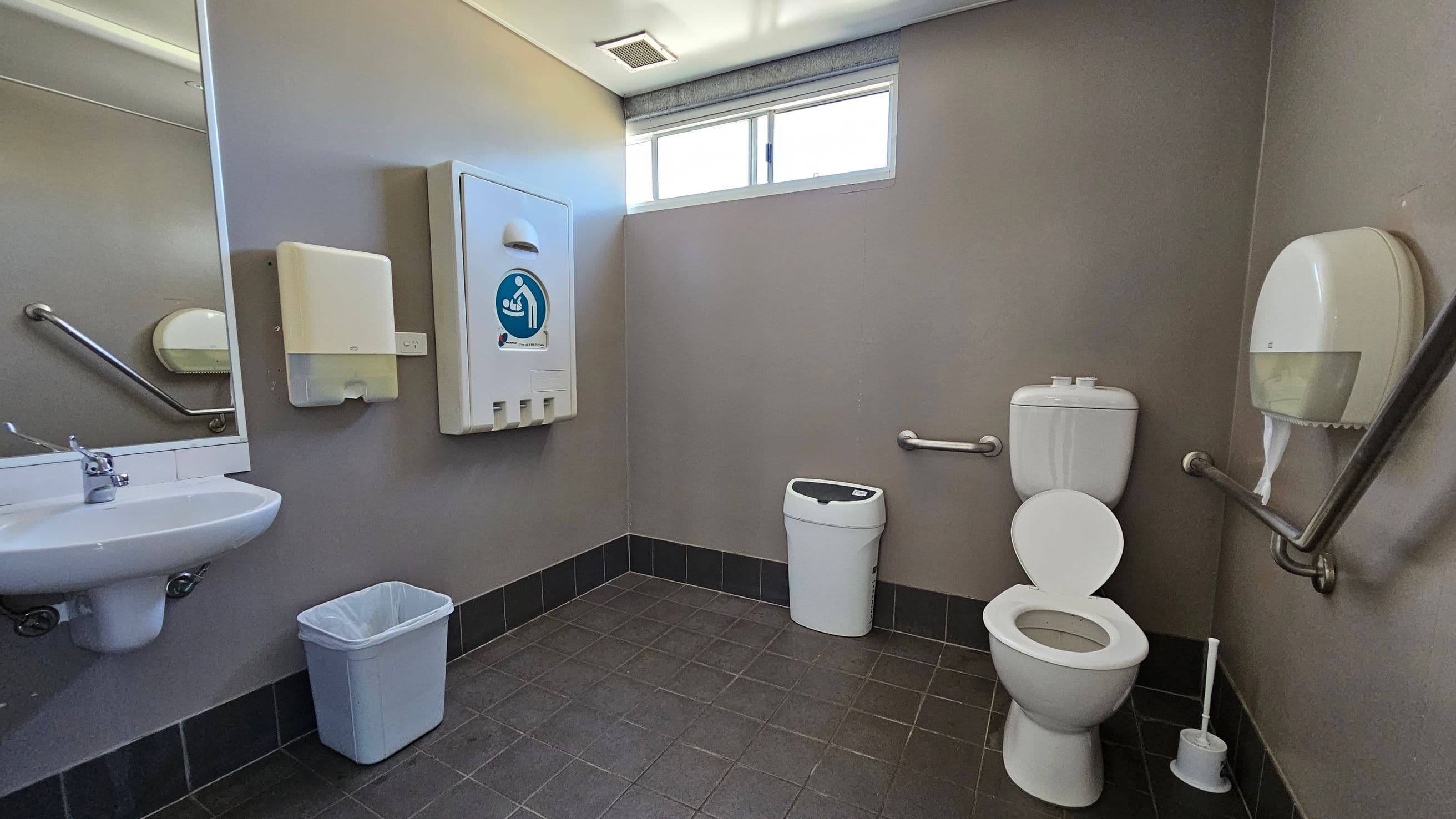A public toilet with grey walls and a tiled floor. On the left, there's a white sink with a mirror above it and a paper towel dispenser. Below the sink is a blue waste bin. In the centre, a baby changing station is mounted on the wall. On the right, a white toilet with grab bars on either side and a toilet brush nearby. Above the toilet is a small window, and a toilet paper dispenser is mounted on the wall.