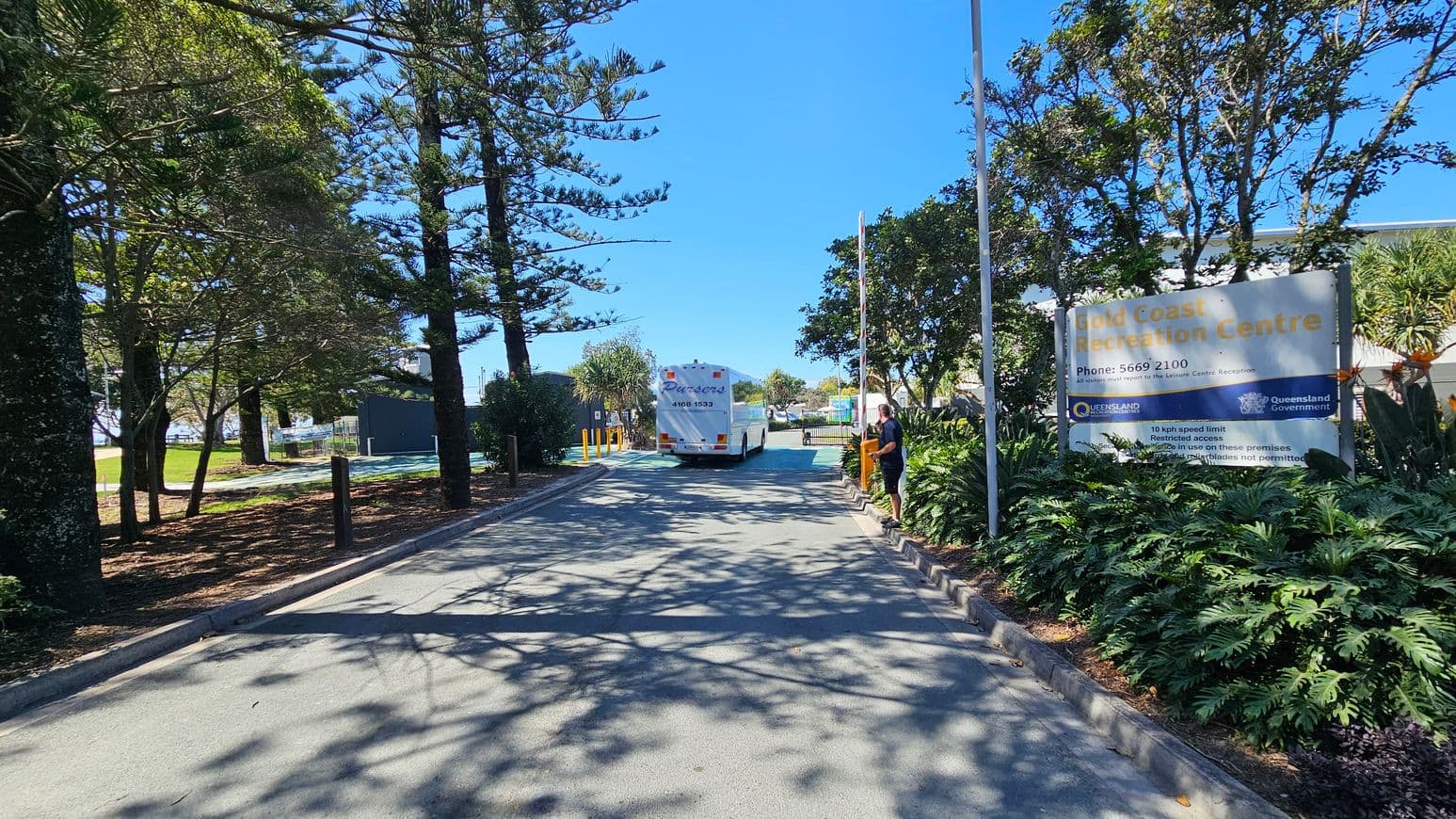 A road lined with tall trees on the left, casting shadows on the pavement. On the right, a large sign reads "Gold Coast Recreation Centre" with contact information and rules. A person stands near the sign. In the centre, a white bus is driving away, with greenery and more trees in the background. The scene is well-lit, suggesting a sunny day.