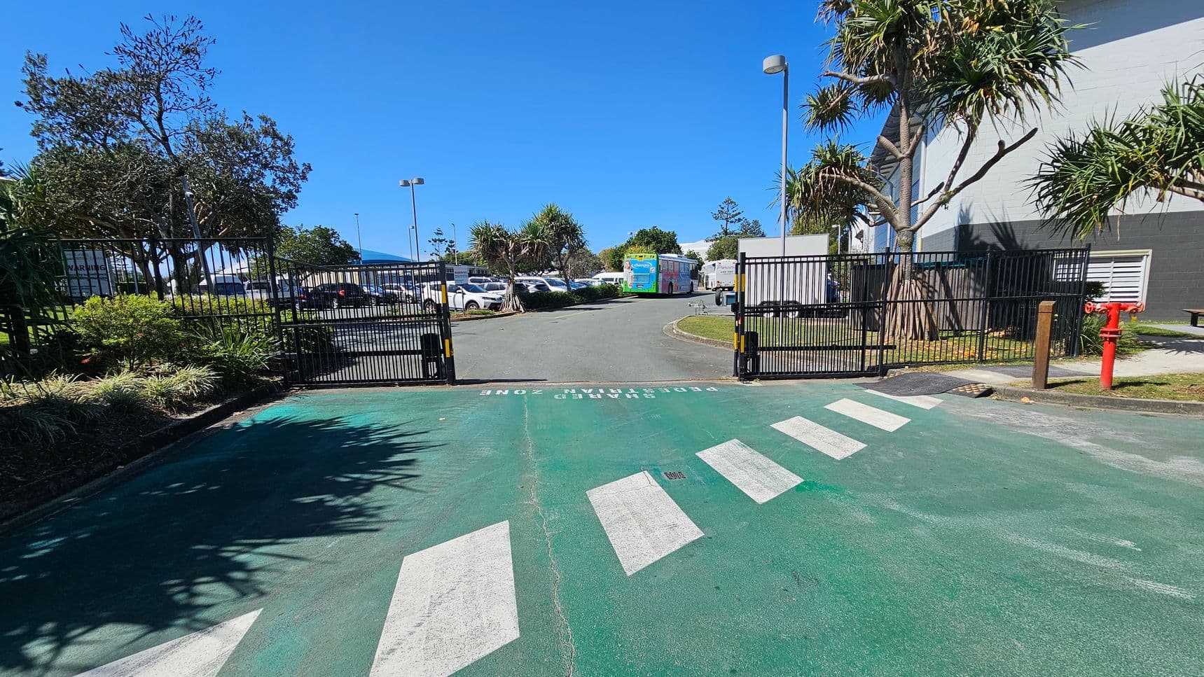 A gated entrance with a green pedestrian zone and white zebra crossing in the foreground. The gate is open, leading to a car park with several vehicles and a colourful bus in the background. On the left, there are trees and shrubs, while on the right, a building with a grey and white exterior and a red fire hydrant is visible. The sky is clear and blue.