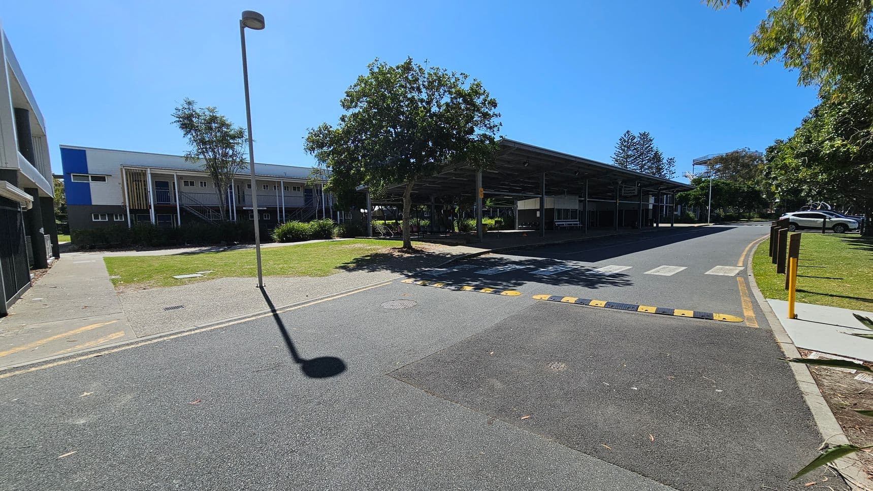 A paved road with a speed bump and a pedestrian crossing runs through the centre. On the left, there's a building with a blue and white facade, and a grassy area with a tree. On the right, a covered structure with benches and another tree is visible. A car is parked further down the road. The area is surrounded by greenery, and a tall streetlamp casts a shadow on the road.