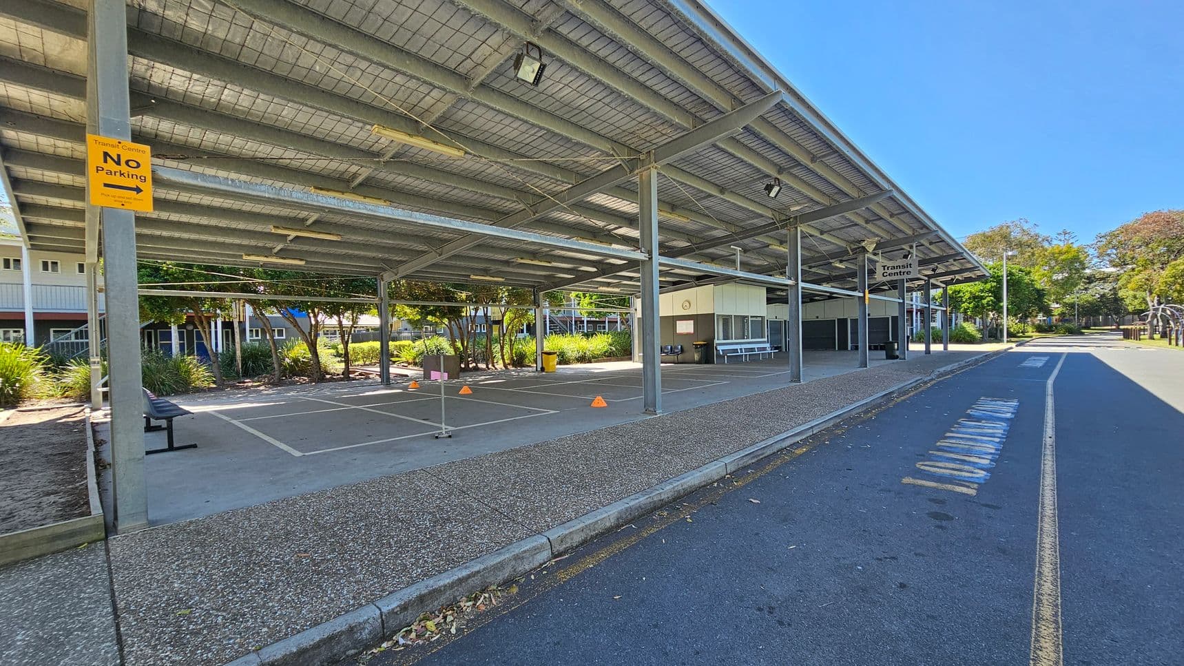 A covered area with a metal roof and support beams, featuring a "No Parking" sign on the left. The ground is paved with a speckled texture. There are marked parking spaces with orange cones and a bench. In the background, there are buildings with windows and greenery, including trees and shrubs. On the right, a road with faded markings runs alongside the structure, and a "Transit Centre" sign is visible. The area is well-lit with natural light.