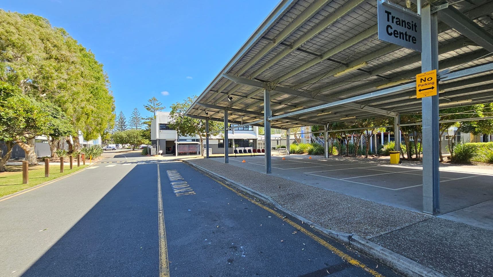 A transit centre with a large metal roof structure on the right, providing shade over a paved area. A sign reads "Transit Centre" with a "No Parking" notice below it. On the left, a road with a marked lane runs alongside a row of trees and grass. In the background, there are buildings with trees and shrubs around them. The ground is a mix of asphalt and concrete, and the area is well-lit.