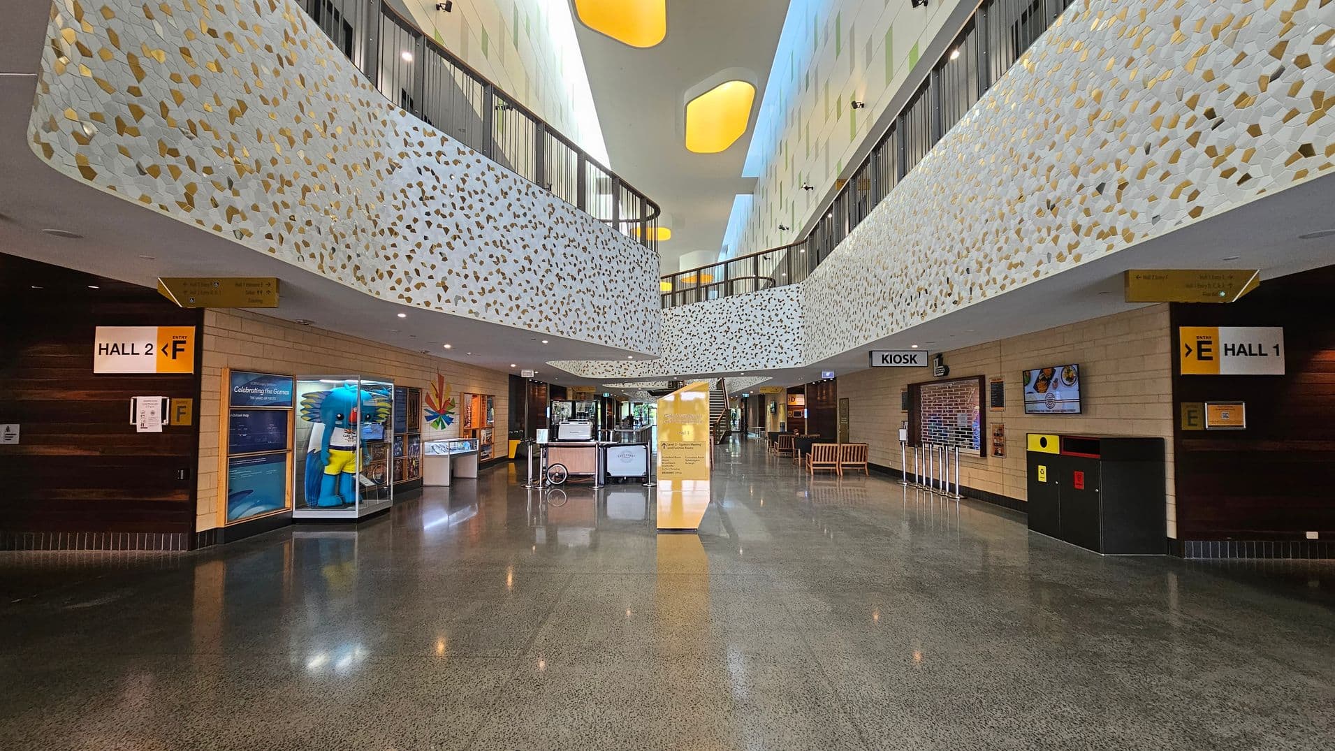 A spacious indoor area with a polished floor. On the left, there is a sign for Hall 2 and a display featuring a large blue mascot. On the right, a sign for Hall 1 and a kiosk are visible. The upper level features a decorative railing with a white and gold pattern. In the centre, there is a yellow information stand and a staircase leading upwards. The ceiling has several recessed, oval-shaped lights.