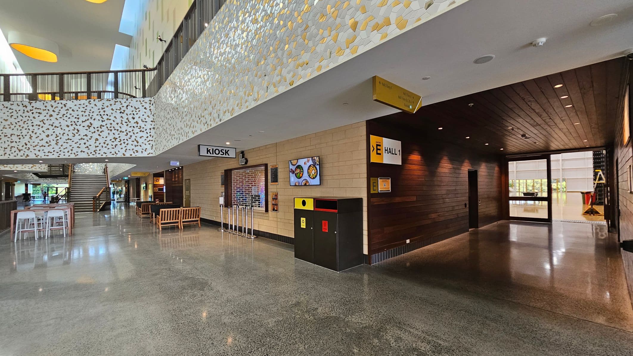 A spacious indoor area with a polished concrete floor. On the left, there are high tables with stools and a staircase leading to an upper level. The ceiling has geometric cut-outs with yellow accents. On the right, a kiosk area with a sign, a digital screen displaying food images, and a waste bin. The wall is decorated with a mosaic pattern. In the background, large windows allow natural light to enter, and there are wooden benches and a dark wood-panelled hallway.