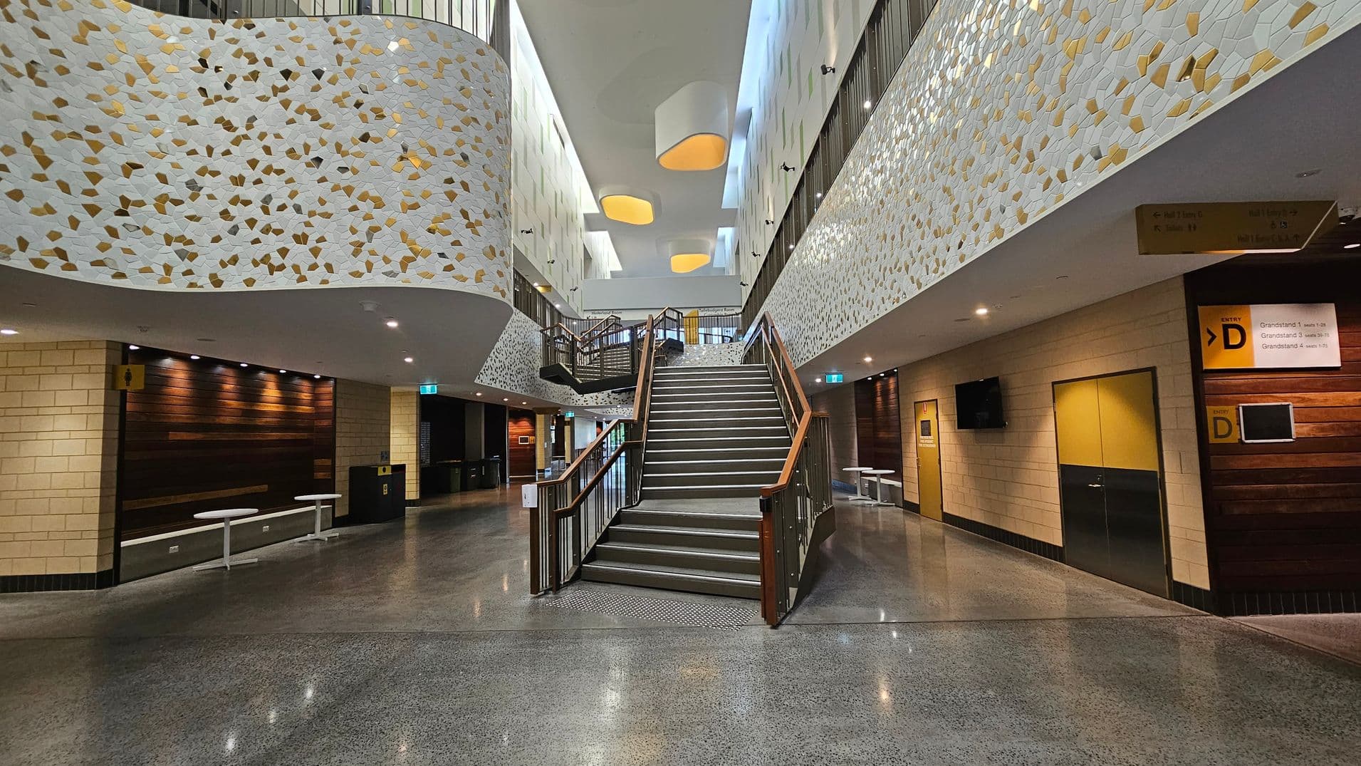 A spacious interior with a central staircase leading upwards. The walls are adorned with a decorative pattern of white and gold shapes. On the left, there are wooden panels and a seating area with white tables. On the right, there are yellow and black doors with signage above them. The floor is polished concrete, and the ceiling features large, geometric light fixtures. The area is well-lit with overhead lights.