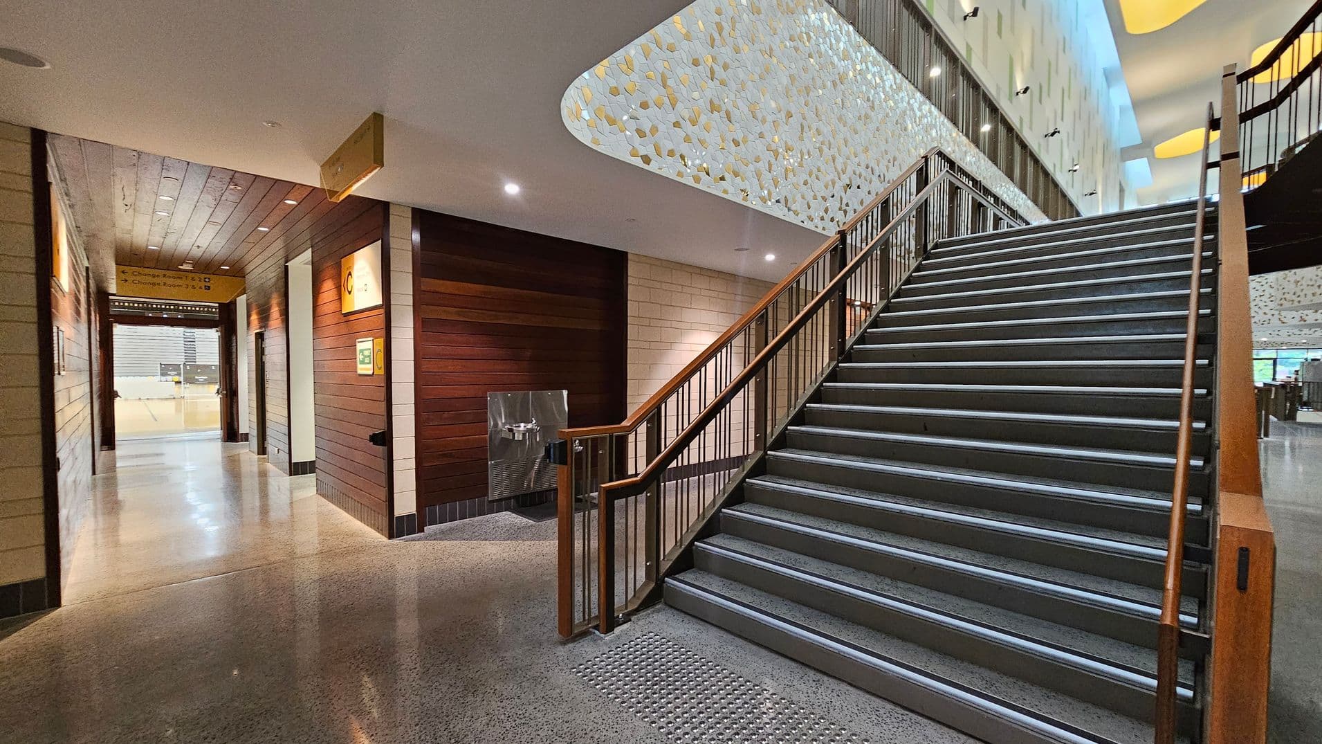 A modern indoor space with a wide staircase on the right, featuring wooden handrails and metal steps. The left side has a corridor with wooden panelled walls and a polished concrete floor, leading to a distant area. Above, a ceiling with a decorative pattern of light-coloured tiles. A water fountain is positioned near the base of the stairs. The area is well-lit with recessed lighting.