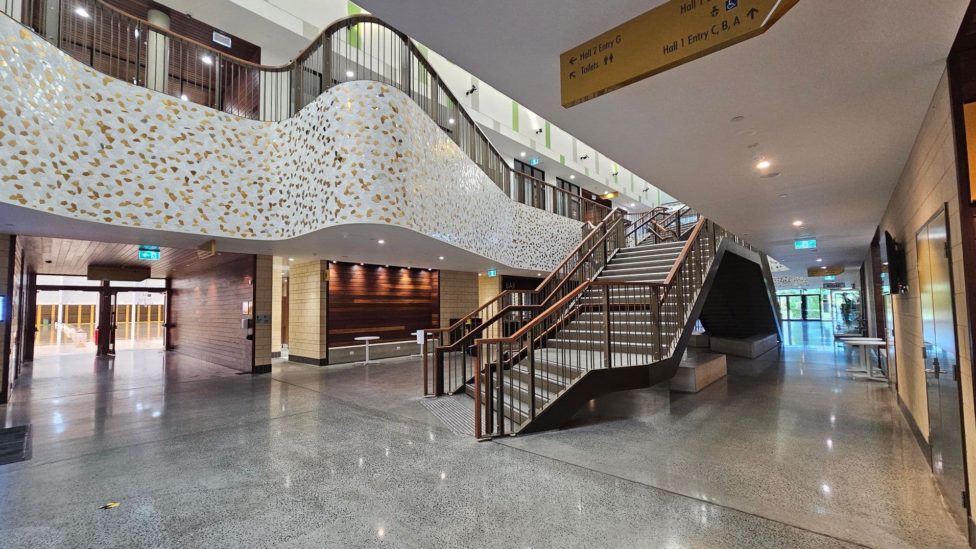 A modern interior with a polished concrete floor and a white ceiling. On the left, there's a curved wall with a terrazzo pattern in white and gold. Below it, a wooden wall with spotlights and a small seating area with white tables. In the centre, a wide staircase with wooden handrails leads to an upper level. On the right, a corridor with glass doors at the end. Overhead, a yellow sign indicates directions to various halls and facilities.