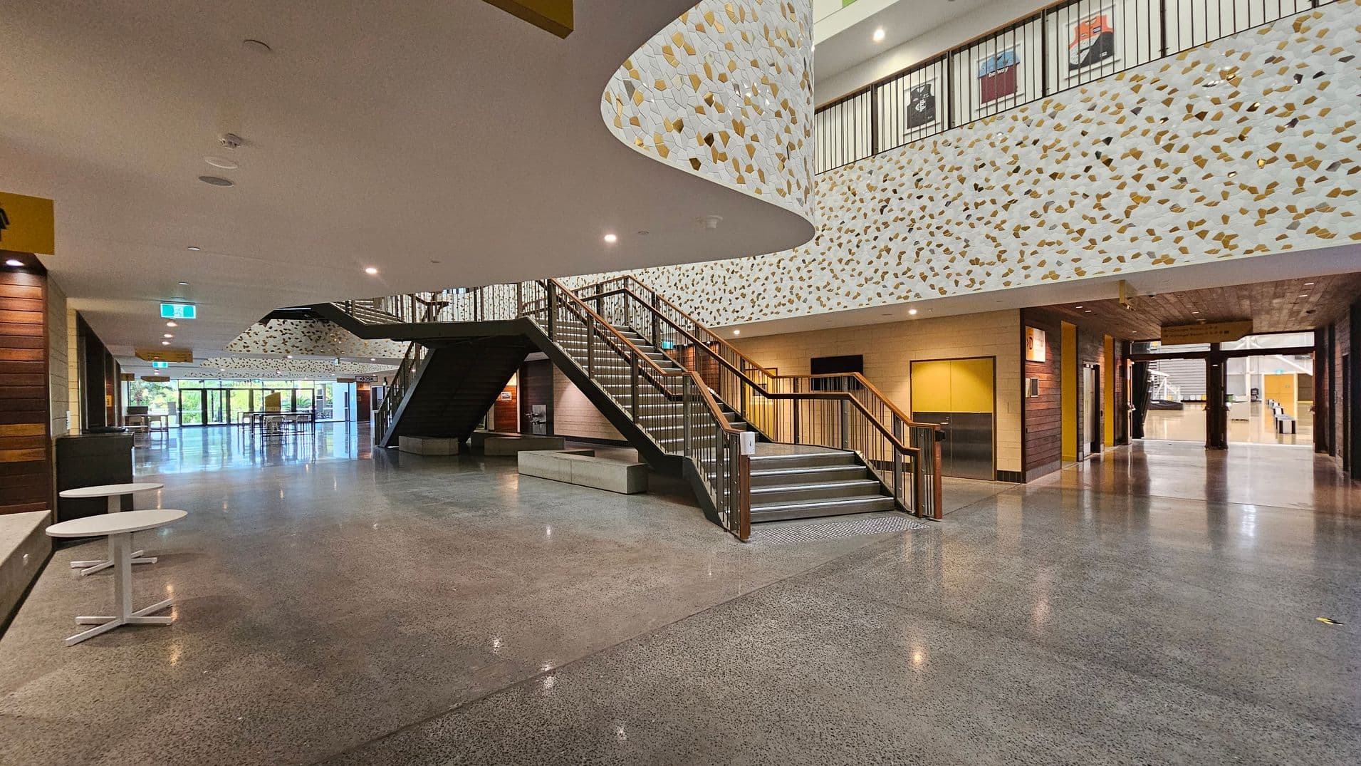 A spacious, modern interior with polished concrete floors and a high ceiling. On the left, there are small white tables and a wooden wall. In the centre, a wide staircase with wooden railings leads to an upper level. The walls are decorated with a geometric pattern in white, gold, and brown. On the right, there are entrances to other rooms and a corridor. Large glass doors at the far end let in natural light, revealing greenery outside.