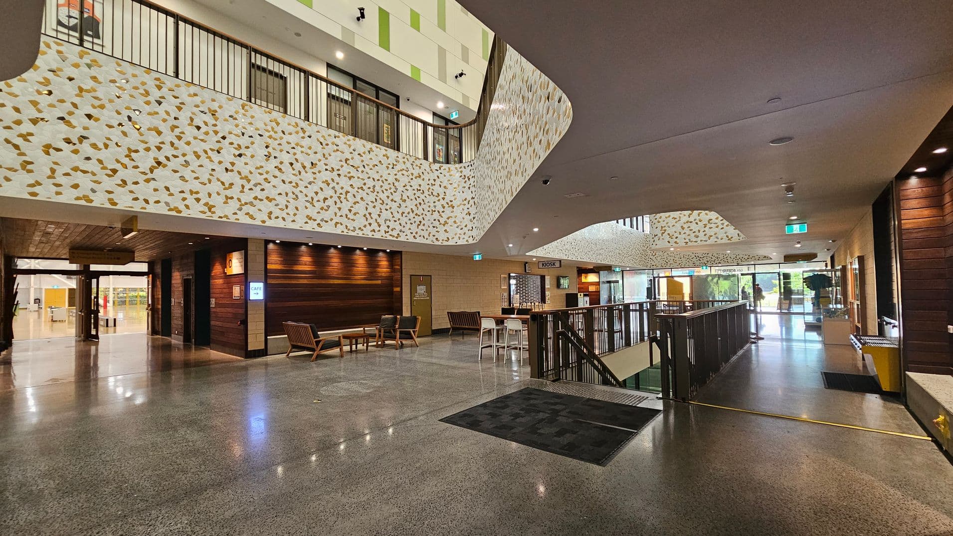 Spacious interior with polished concrete floors and a high ceiling. On the left, there's a seating area with wooden chairs and a table against a wall with wooden panelling. Above, a balcony with a decorative railing featuring a pattern of gold and white. On the right, a kiosk area with stools and a counter. Large windows in the background allow natural light to illuminate the space. An escalator is visible in the centre, leading to a lower level.