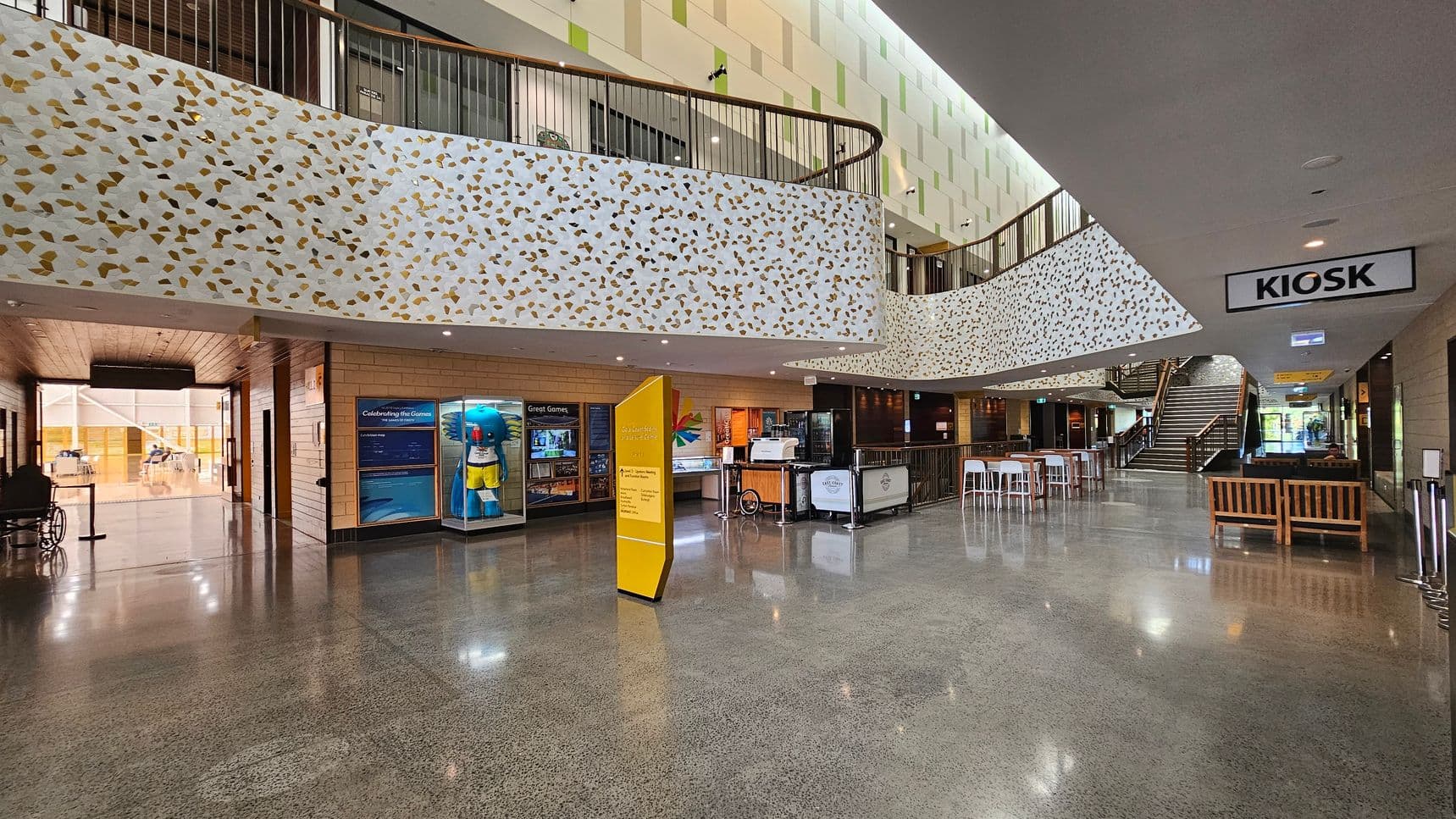 A spacious indoor area with a polished concrete floor. On the left, there is a display with a large blue mascot and informational panels. In the centre, a yellow sign stands near a kiosk with a counter and stools. The upper level features a decorative railing with a terrazzo pattern. On the right, wooden benches are placed near a staircase leading to the upper floor. The ceiling is smooth and grey, with recessed lighting.