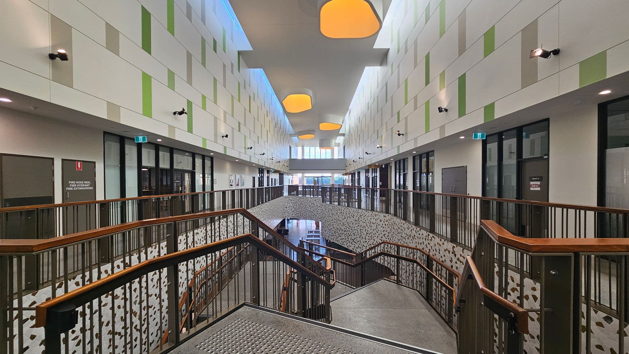 A modern interior with a wide staircase in the centre, featuring wooden handrails and patterned side panels. The walls are decorated with a geometric pattern in shades of green and beige. On both sides, there are glass doors and windows leading to various rooms. The ceiling is high with large, round, recessed lights. The floor is grey and polished, and there are emergency exit signs above some doors.