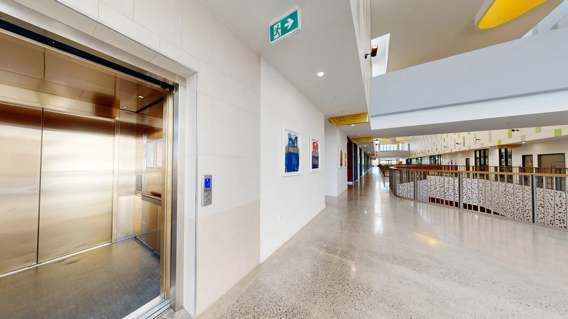 A modern indoor corridor with a polished concrete floor. On the left, an open lift with metallic doors. The right side features a long hallway with a railing overlooking a lower level. The walls are adorned with framed pictures. Above, a green exit sign is visible. The ceiling has recessed lighting and some yellow accents. The corridor extends into the distance with large windows at the far end.