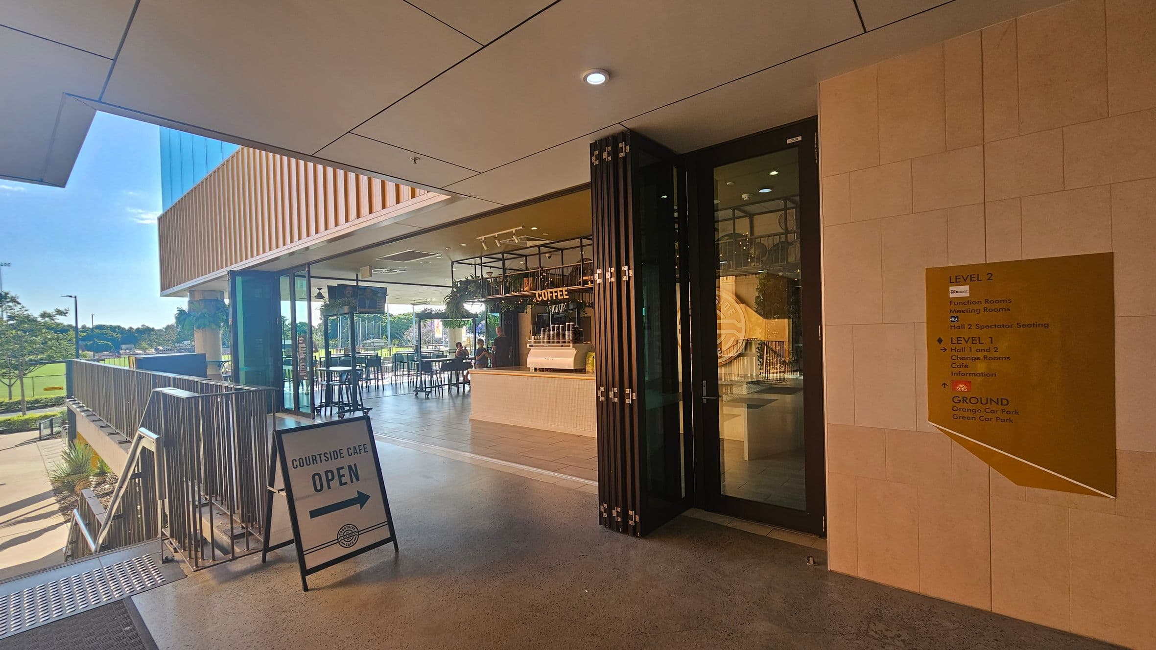 A modern café entrance with a sign reading "Courtside Café Open" on the left, pointing towards the café. The café interior features a counter with a "Coffee Pick Up" sign and several tables and chairs. The exterior has a glass façade and a wooden panelled overhang. A directory sign on the right lists different levels and facilities. The floor is polished concrete, and the ceiling has recessed lighting. Outside, there is a view of a green field and trees.