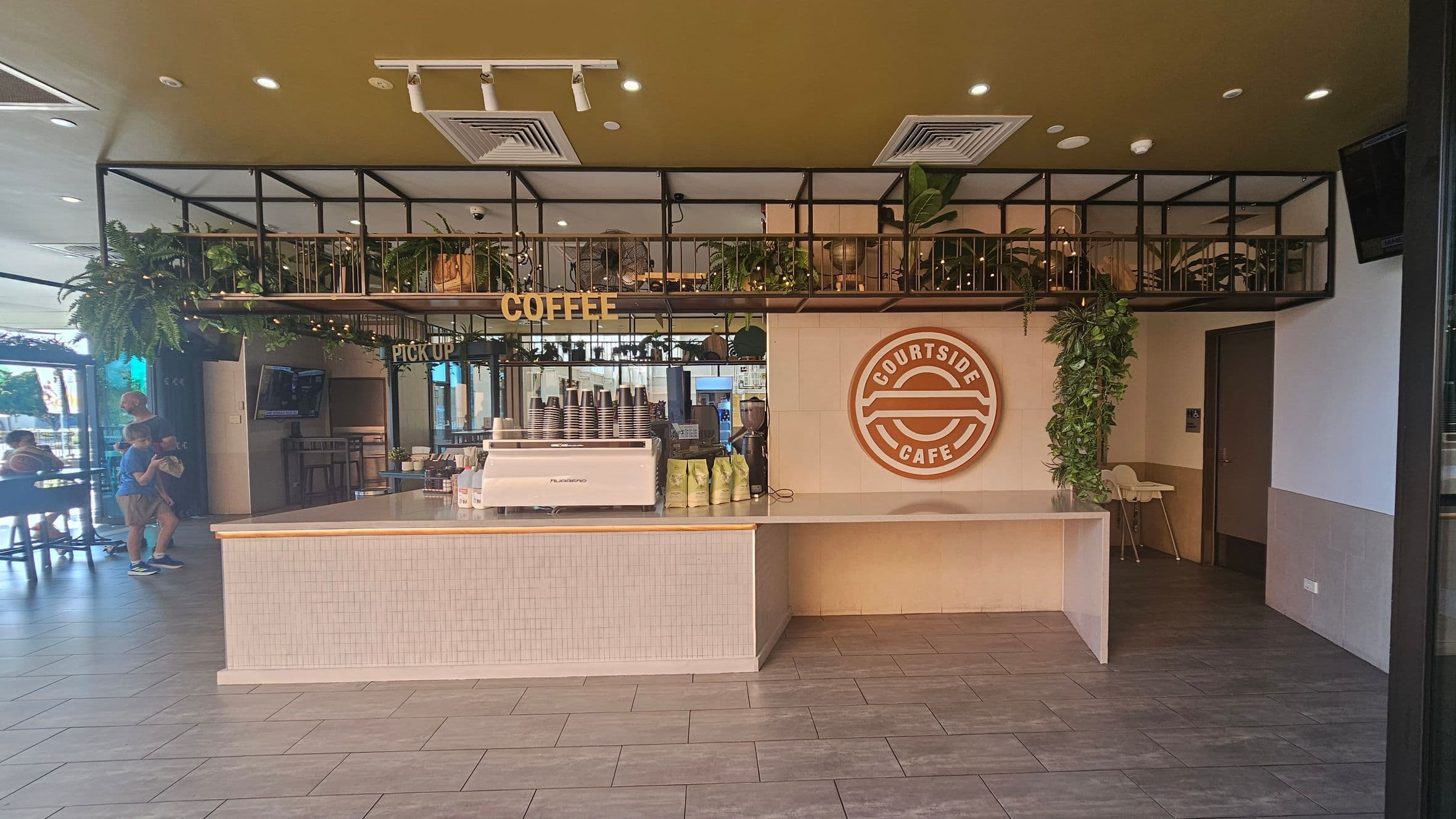 A café interior with a counter in the centre featuring a coffee machine and cups. Above the counter, a sign reads "COFFEE" with plants and decorative items on a shelf. The wall behind has a large "COURTSIDE CAFE" logo. To the left, a seating area with people visible through glass doors. The floor is tiled, and the ceiling has recessed lighting and air vents. Greenery hangs from the upper shelves, adding a natural touch.