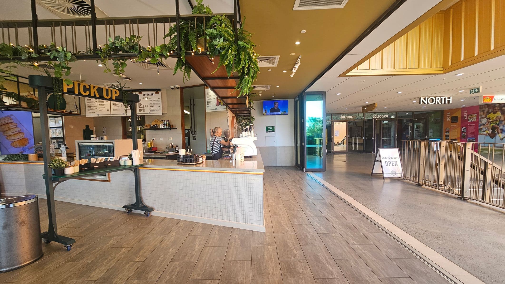 A café interior with a counter on the left, featuring a "PICK UP" sign and a barista preparing drinks. The counter is adorned with plants and a menu board. The floor is wooden, and the ceiling has recessed lighting. On the right, there's an open area leading to a corridor with a sign reading "NORTH" and a "COURTSIDE GATE OPEN" sign. The corridor has glass doors and posters on the walls.