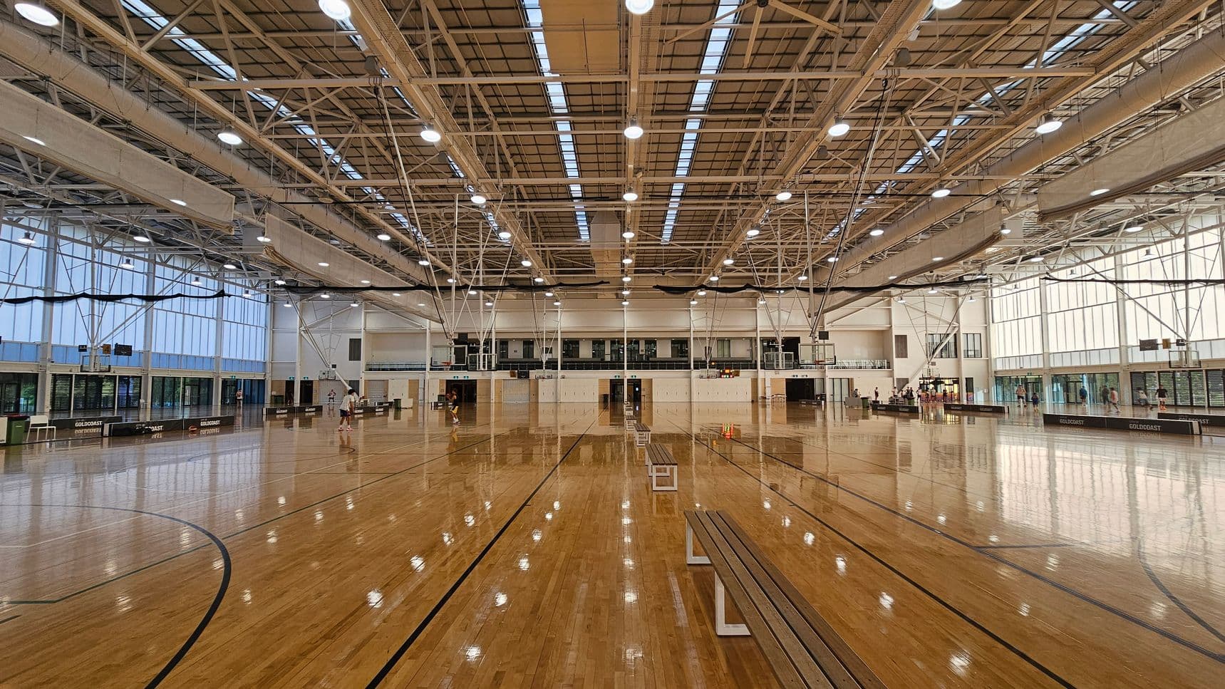 A large indoor sports hall with a polished wooden floor and high ceiling featuring exposed beams and bright lighting. The left and right sides have tall windows allowing natural light. In the centre, there are multiple basketball courts with hoops. Benches are placed along the courts, and a few people are visible in the background, possibly engaging in sports activities. The walls are lined with banners and equipment.