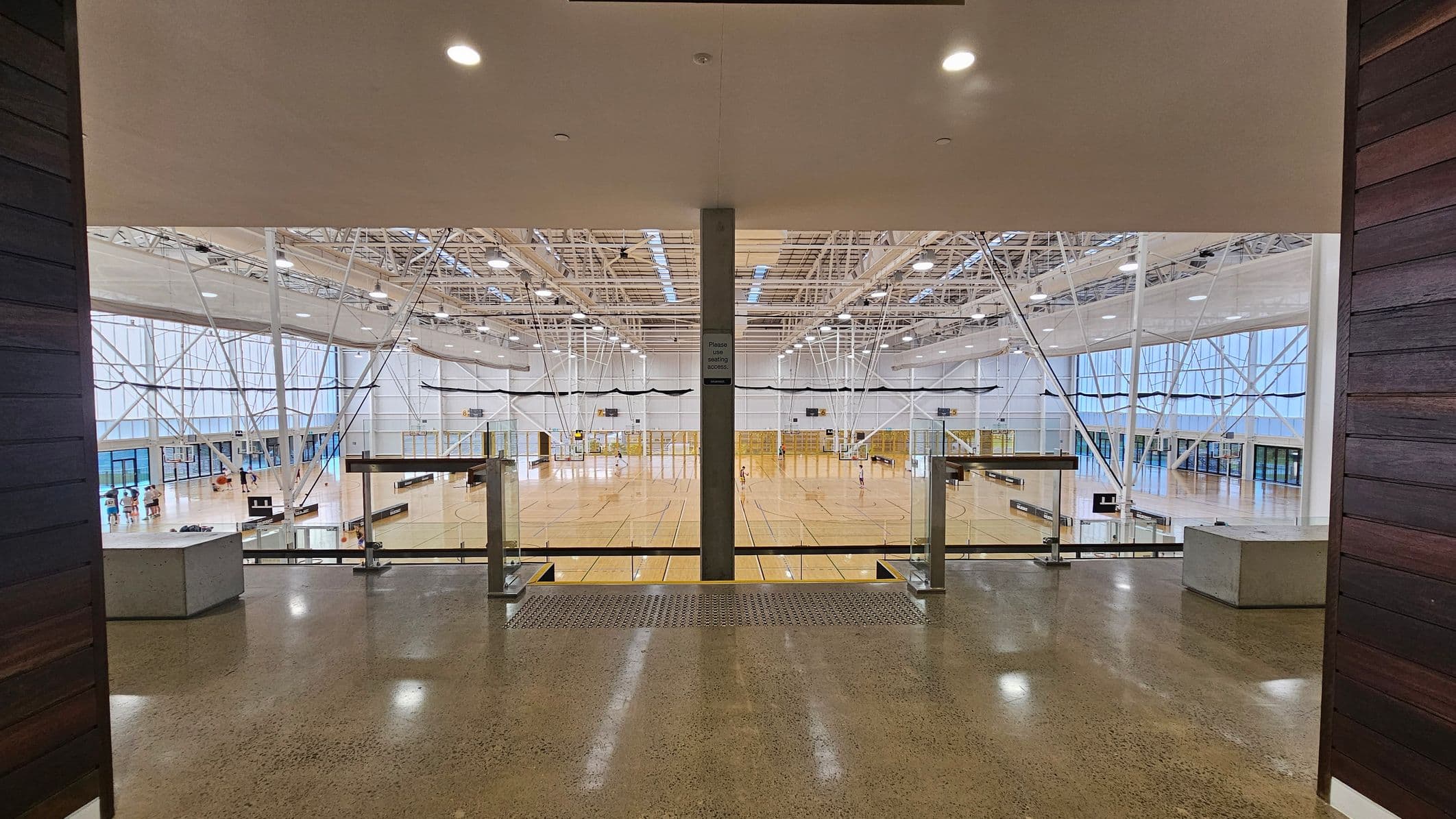 A large indoor sports hall with a polished wooden floor and high ceiling. The hall is divided into multiple courts with visible basketball hoops and netting. The ceiling features exposed beams and numerous lights. On the left, there are large windows and a few people near the entrance. The foreground has a polished concrete floor with a metal railing and a central pillar. The walls are lined with glass and metal structures.
