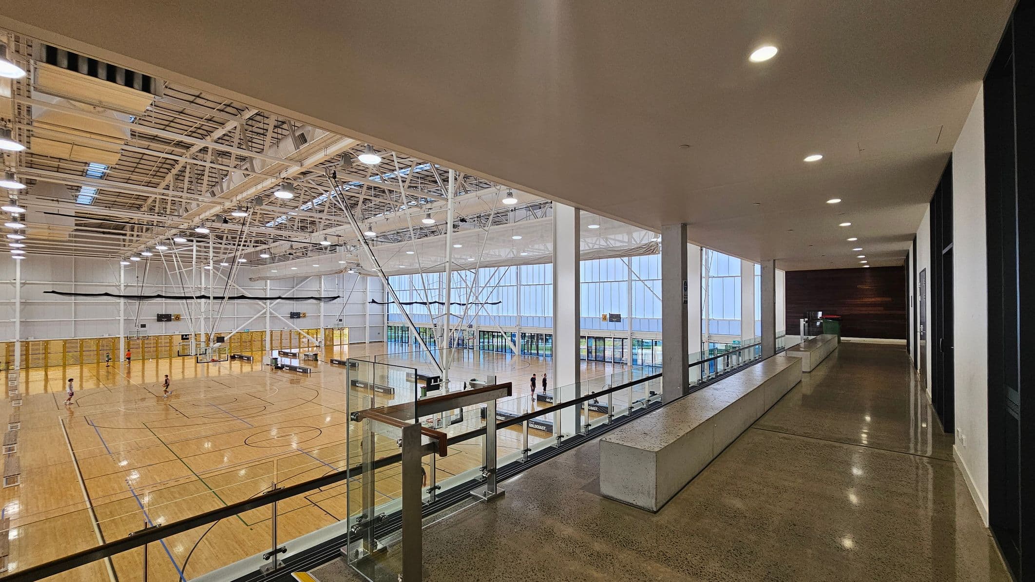 A spacious indoor sports hall with a polished wooden floor and multiple basketball courts. The left side features high ceilings with exposed beams and bright lighting. On the right, a corridor with a polished concrete floor and a long bench runs alongside a glass railing overlooking the courts. Large windows in the background allow natural light to flood the space. A few people are visible on the courts and in the corridor.