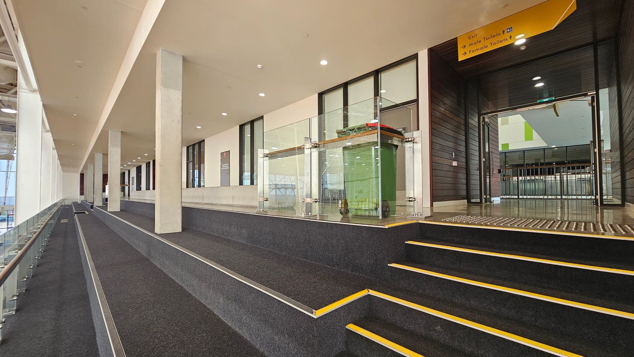 A modern indoor space with a wide, carpeted ramp on the left, bordered by glass railings. Several concrete pillars line the ramp. On the right, there are steps with yellow safety edges leading to a glass-enclosed area containing a green bin. Above, a yellow sign indicates directions to exits and toilets. The ceiling is smooth with recessed lighting, and the floor is dark with a textured surface near the entrance.