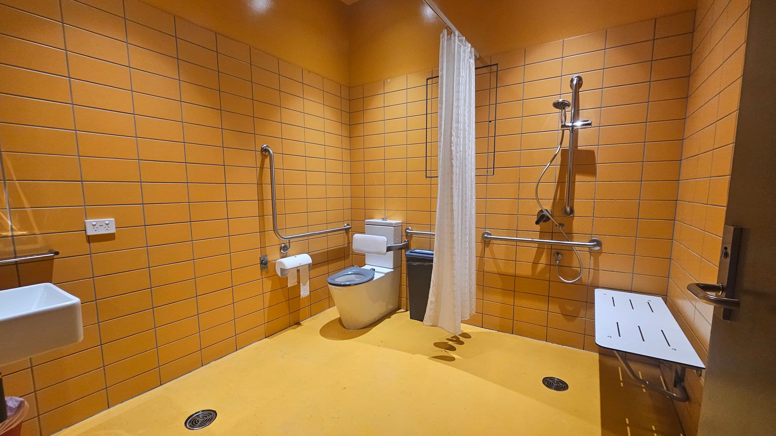 A bathroom with orange tiled walls and a yellow floor. In the centre, there's a toilet with grab bars on both sides and a bin nearby. To the right, a shower area with a handheld showerhead and a foldable bench. A white curtain separates the shower from the rest of the room. On the left, a small white sink with a soap dispenser and a power outlet above it. The ceiling is also orange, matching the walls.