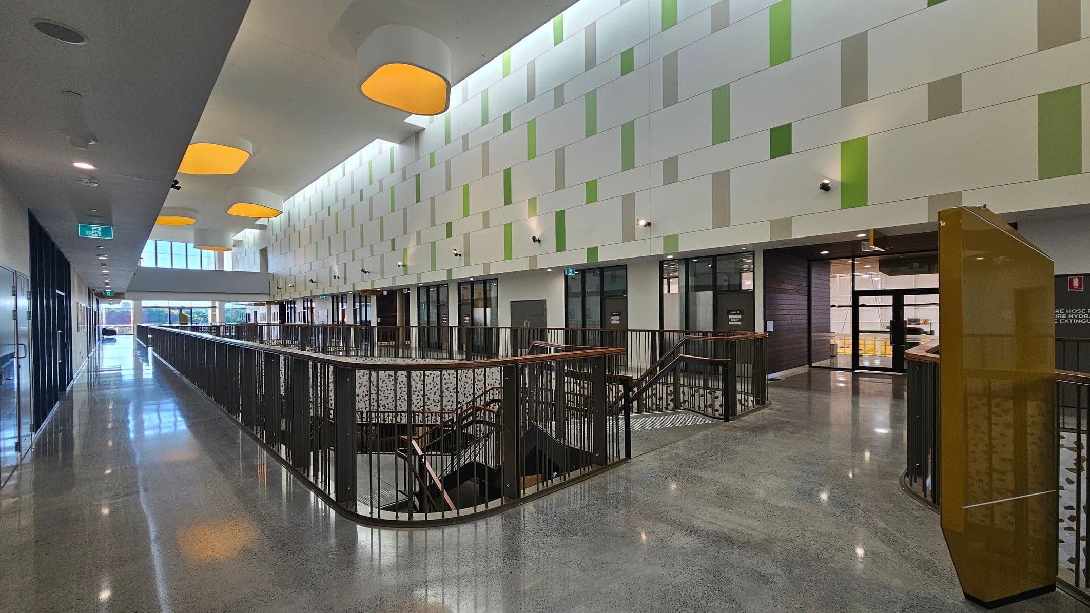 A modern interior with a polished concrete floor and a high ceiling featuring large, recessed yellow lights. On the left, a long corridor with glass doors and windows. The right side has a patterned wall with green and beige panels, and multiple doors leading to rooms. In the centre, a railing surrounds a staircase leading down. The far end of the corridor has large windows allowing natural light to flood in.