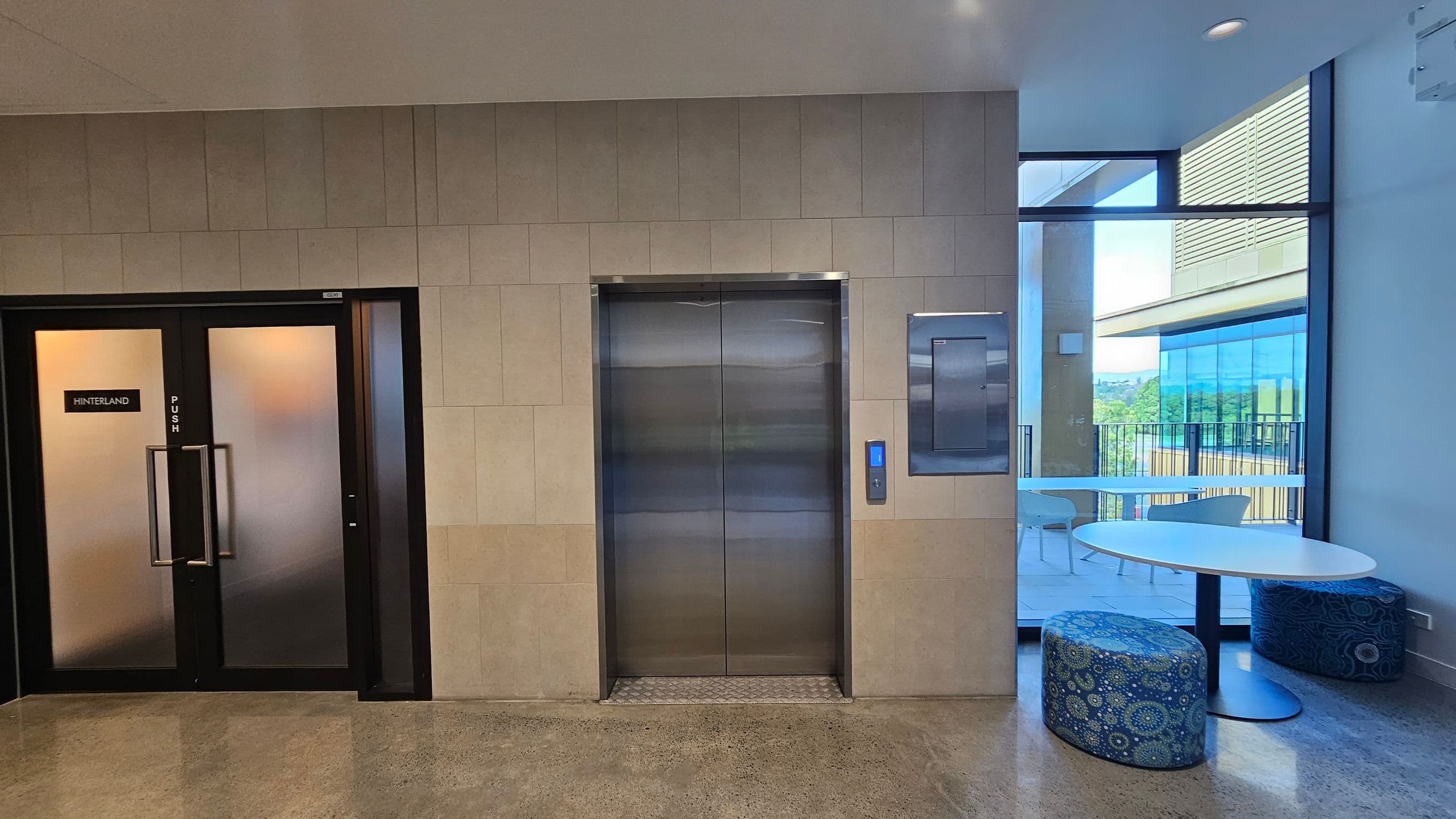 On the left, there are double doors with frosted glass and a sign reading "Hinterland." In the centre, a stainless steel lift with a control panel is set into a tiled wall. On the right, a large window offers a view of a balcony with a table and chairs. Inside, a round white table with patterned blue stools is visible. The floor is polished concrete, and the ceiling is plain with recessed lighting.